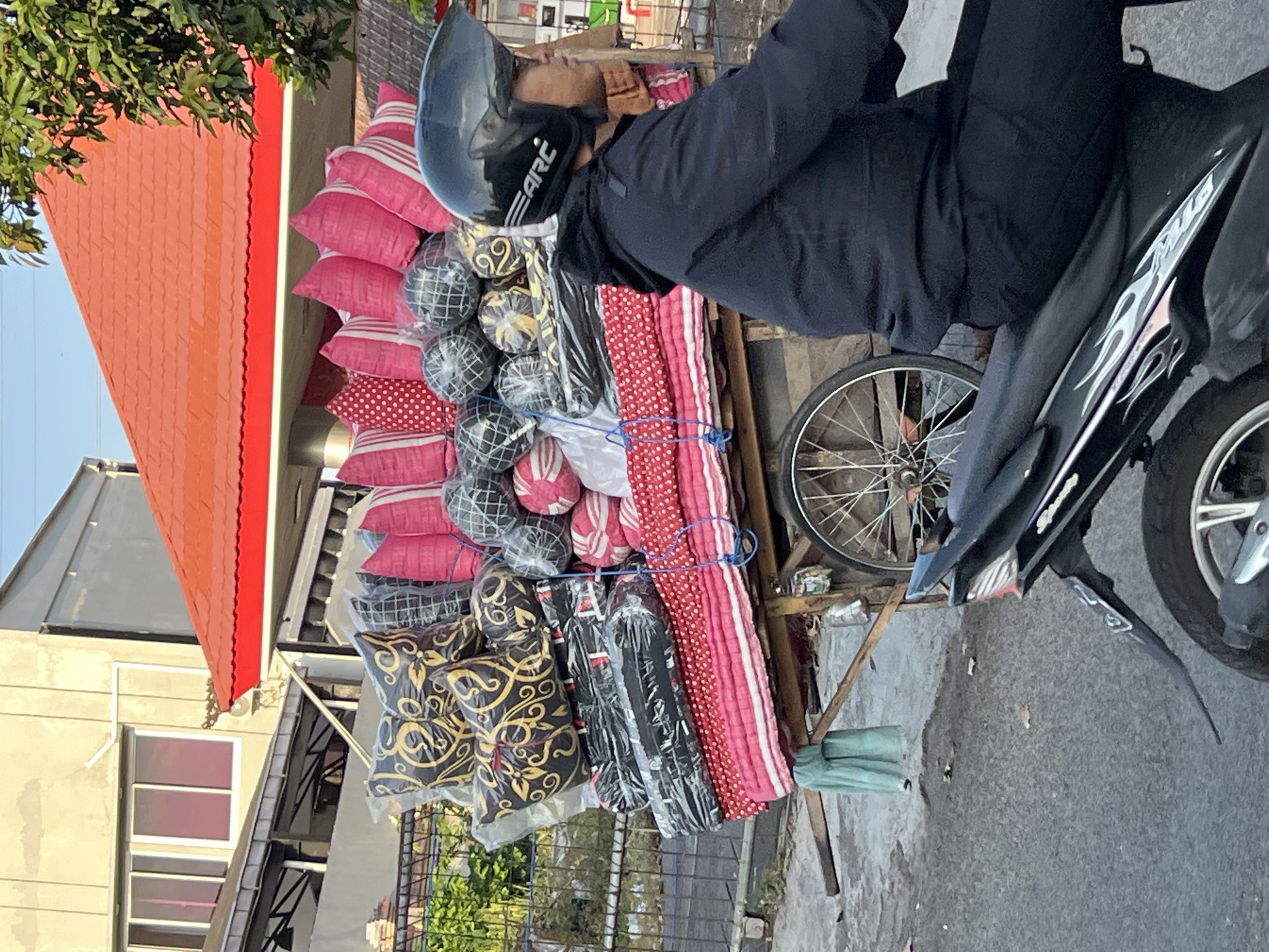 An outdoor, eye-level, medium shot captures a person wearing a black jacket and helmet riding a black motorcycle on a paved street. The person is positioned in the right side of the frame, facing right, with the motorcycle moving towards the right. The motorcycle's rear is visible at the bottom of the frame. In the center of the frame, a wooden cart is laden with an assortment of pillows and rolled bedding. The pillows are in various sizes and patterns, including pink with white polka dots, pink and white stripes, and black with a gold swirling pattern. Some are wrapped in clear plastic. The cart has two spoked wheels, one of which is clearly visible on the right side of the cart. Behind the cart, a building with a red-tiled roof and light-colored walls is visible. The sky is bright and clear, suggesting daylight. The general setting appears to be a street in Denpasar, Indonesia, likely a commercial or residential area given the building and the goods being transported.
