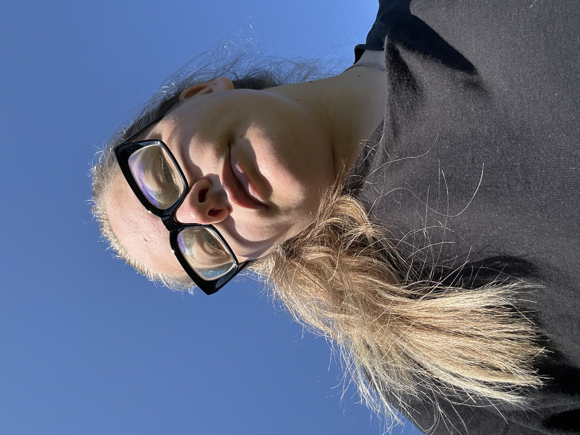 A low-angle, close-up, outdoor shot shows a person from the chest up, facing the camera with a blue sky as the background. The person is wearing black-rimmed glasses and a black t-shirt. Their blonde hair is visible, with some strands falling forward. The lighting suggests a sunny day. The image does not contain any visual information that would indicate the location within the city of Калицино, Russia.