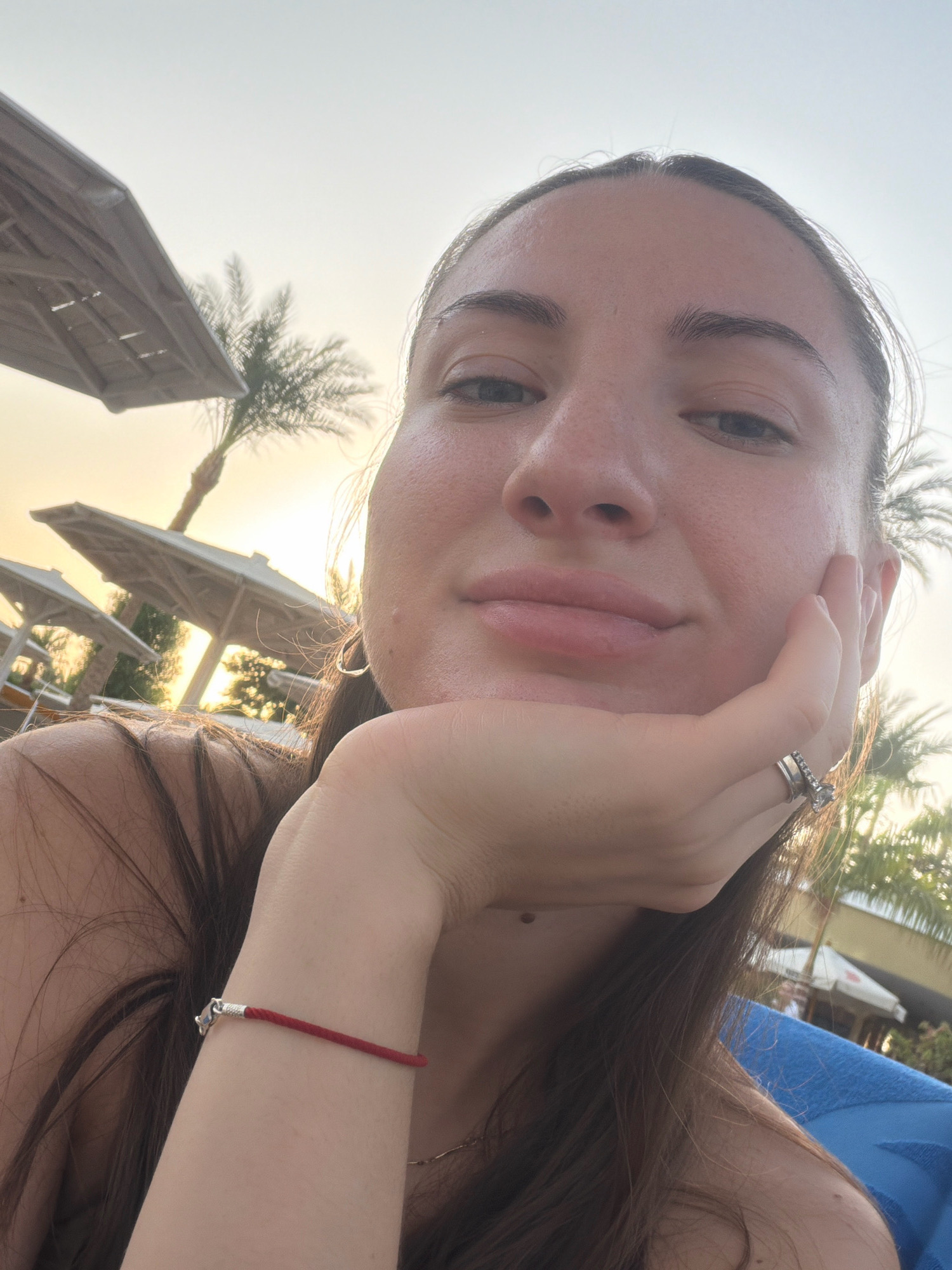 A young woman with long brown hair takes a selfie outdoors. She rests her chin on her hand, wearing a silver ring with stones and a silver bracelet with a red cord. Her left shoulder is visible. In the background, palm trees and beach umbrellas suggest a resort or hotel setting in Sharm El Sheikh, Egypt, with the sun setting or rising, casting a warm glow.