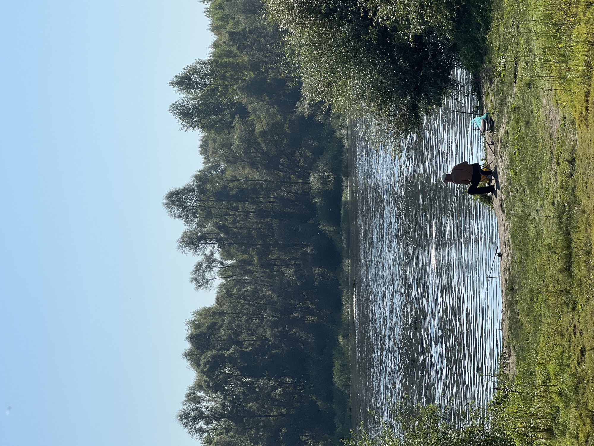 In Salavat, Russia, a person is fishing by the river on a sunny day. The person is seated on a yellow foldable chair on the riverbank, holding a fishing rod and casting a line into the water. A blue bag and some other fishing equipment are placed near the chair. The river is calm with gentle ripples, reflecting the clear blue sky and the dense green trees lining the opposite bank. The foreground features a grassy area with patches of dirt. The scene captures a tranquil moment of recreational fishing amidst a natural setting.