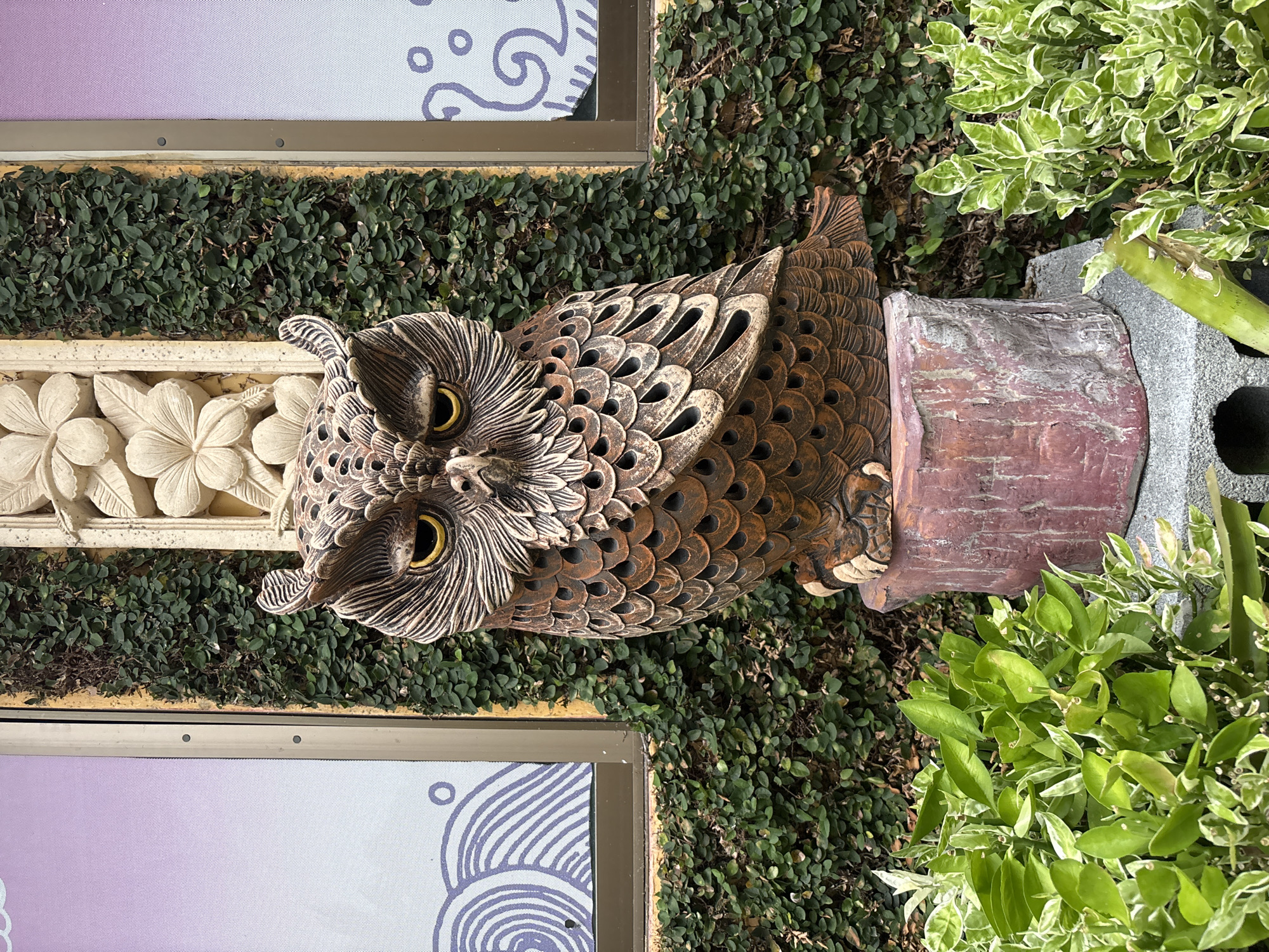 An owl statue made of weathered, brown, textured material sits on a reddish-pink stone pedestal in front of a wall covered in dense green ivy. The owl has large, yellow eyes and intricate detailing on its feathers and face, with cut-out sections creating a hollow effect. To the left and right of the owl are windows with a purple gradient and stylized white wave patterns. Above the owl, a section of decorative carved stone with floral motifs is visible against a yellow wall. In the foreground, lush green plants with variegated leaves surround the base of the statue. The scene appears to be outdoors, likely in a garden or courtyard setting in Chalong, Thailand.