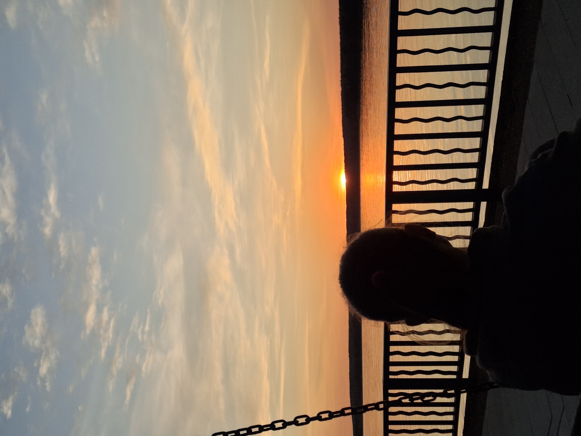 The image shows a sunset over a body of water, likely a river or lake, as viewed from a dock or pier in Chaikovsky, Russia. The sun is setting directly on the horizon, creating a bright orange glow that is reflected on the water's surface. The sky is a mix of orange, yellow, and blue hues. A silhouette of a person and the dock railings are visible in the foreground.