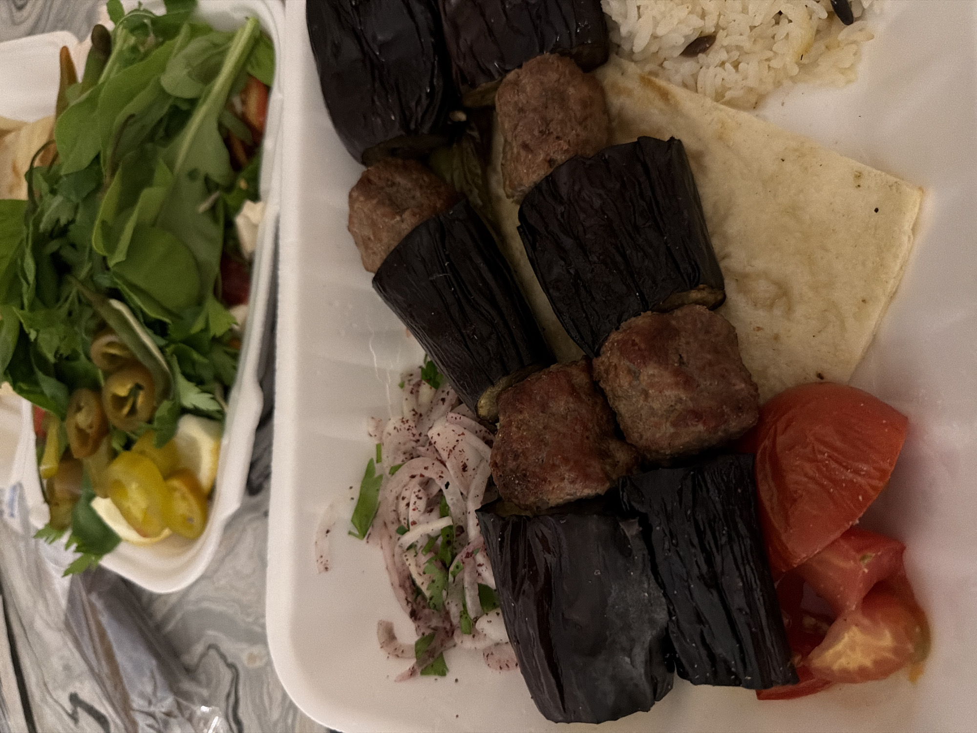 An overhead shot displays a takeaway meal on a marble-patterned surface. The meal is divided into two white rectangular styrofoam containers. The top container holds a salad featuring green leafy vegetables, sliced jalapeños, and pieces of lemon. Below this, the second container is filled with a main dish and sides. The main dish consists of grilled eggplant pieces interspersed with meatballs. To the left of the main dish is a side of sliced red onions sprinkled with sumac and chopped parsley. The bottom right of the container shows a portion of fluffy white rice with dark specks, a piece of flatbread, and sliced tomatoes.