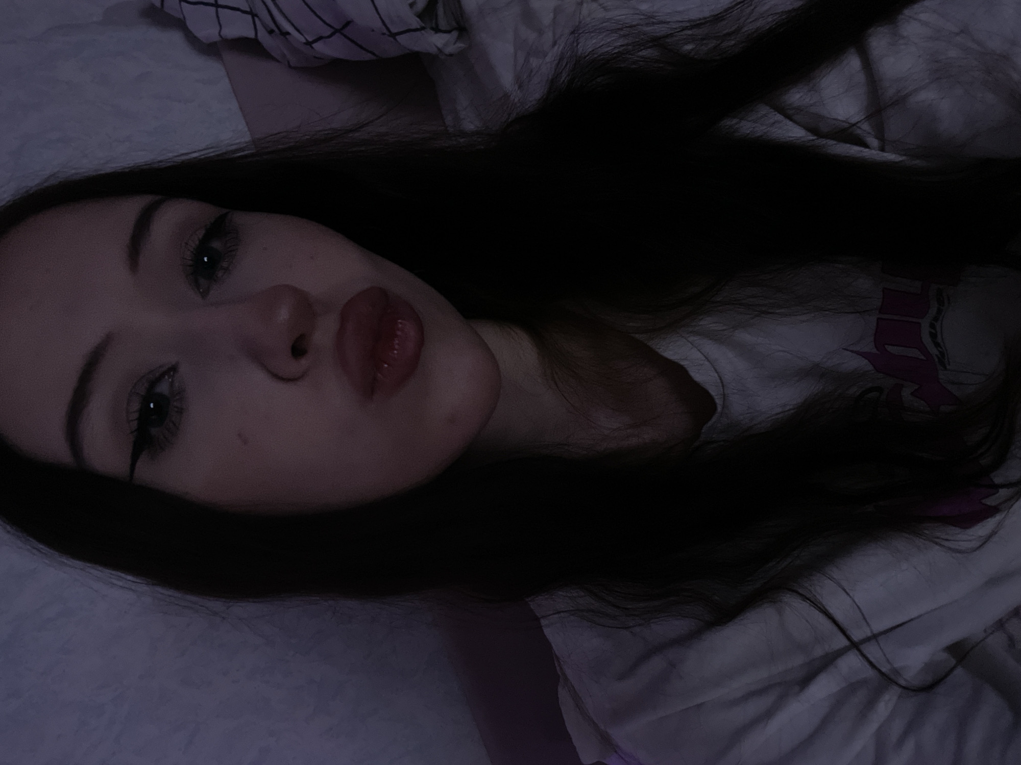 The image is a close-up selfie of a young woman with long, dark hair, taken indoors. She has her lips pursed, and is wearing makeup, including winged eyeliner. She is wearing a white t-shirt. The background appears to be a bed with a textured headboard. The lighting is dim, with a purple hue. No specific location in Votkinsk, Russia, can be determined from the image.
