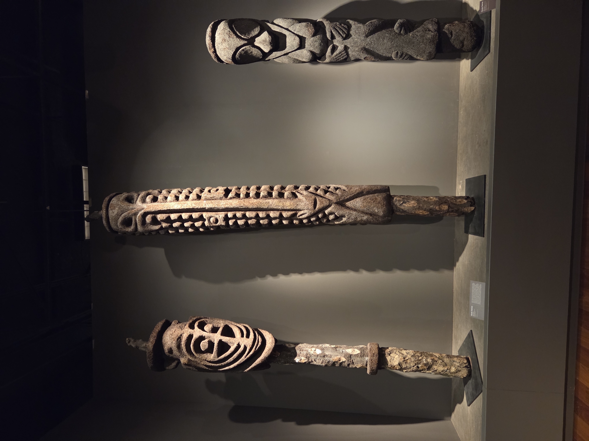 The image displays three stone carvings of ancestral figures, likely from a tribal culture, exhibited in a museum. The carvings are arranged from left to right, with varying heights and stylistic features. The first carving on the left has a distinct face with circular eyes and intricate line work, atop a textured wooden shaft. The central carving is the tallest and features a long, narrow face with a textured pattern of dots and lines running down its length, also with a wooden base. The third carving on the right is a more solid, humanoid figure with broad shoulders and hands clasped at the waist, with a rounded head and stylized features. All three figures are displayed on dark rectangular bases. The background is a plain, dark gray wall, illuminated by spotlighting that casts dramatic shadows. The exhibition space appears to be a museum or gallery, with a polished floor in the foreground. The overall lighting is dim, emphasizing the artifacts. The objects are positioned on a grey, elevated platform. There is a small descriptive plaque in front of the middle statue. This scene is located in the Bowers Museum, Santa Ana, United States of America.