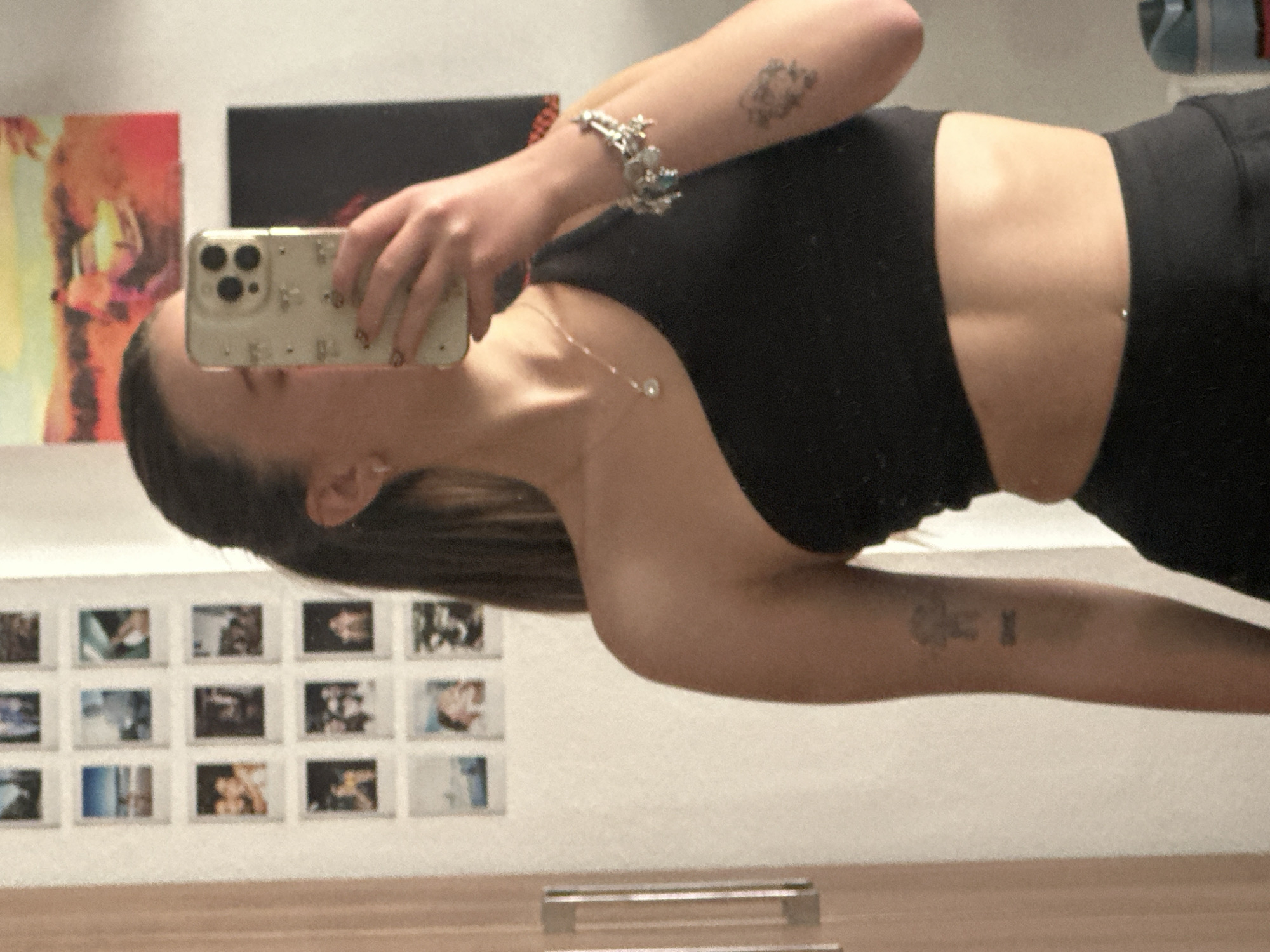 A woman is taking a selfie in what appears to be a public place with artwork on the walls. She is wearing a black sports bra and leggings. She is holding an iPhone with a decorative case in her left hand. She has a silver bracelet on her left wrist and a silver necklace around her neck. She has tattoos on her arms. The artwork on the wall includes colorful abstract pieces and smaller photographs. It is not possible to confirm the specific location in Huixquilucan de Degollado, Mexico.