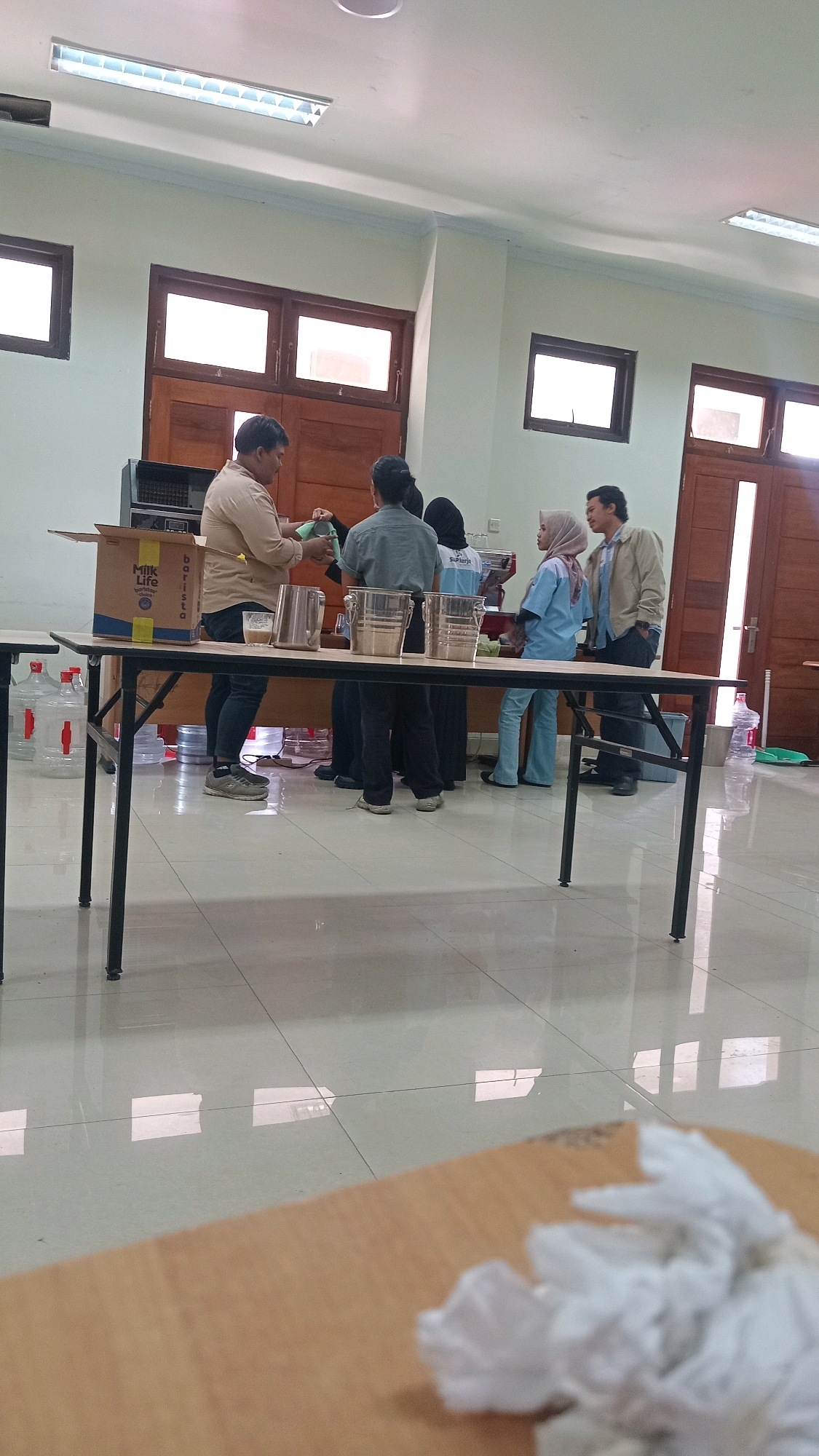 In the city of Lenek, Indonesia, a group of individuals is gathered in what appears to be an indoor community or educational space. The room has pale green walls, a tiled floor with reflections from ceiling lights, and large wooden doors with glass panels, suggesting a modern institutional building.
In the foreground, a long, dark-legged table is positioned horizontally. On the table, there are several large plastic water jugs, and a cardboard box with the "Milk Life" logo is visible. A man in a light brown shirt and dark pants is pouring liquid from a metallic pitcher into a glass. Another person, wearing a gray shirt, stands beside him, and a third person, wearing a black headscarf, is partially visible behind them. Further down the table, two metallic containers with handles are placed, and beyond them, a young woman in a light blue uniform and hijab is interacting with a man wearing a light brown jacket and dark pants. Another person in a blue uniform is also present. In the background, a coffee machine is visible, and a large window offers a glimpse of the outdoors. The scene suggests an event or a demonstration, possibly related to food or beverage service.