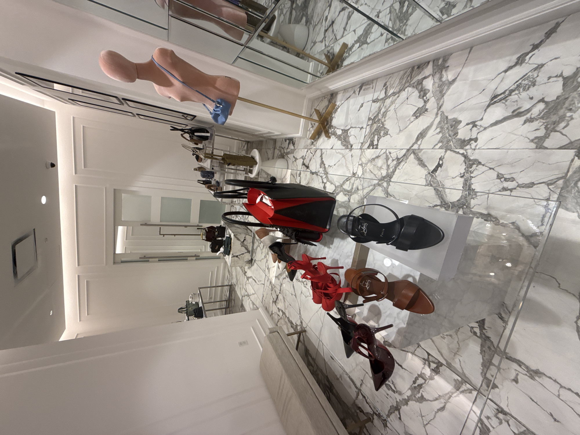 The image depicts the interior of a retail store in Puebla, Mexico, featuring marble flooring and display cases. A mannequin torso holds a small blue purse. A display table showcases various shoes, including black flats, red high heels, and tan sandals, as well as a red and black handbag. The store has recessed lighting and a long hallway with other displays visible in the background.