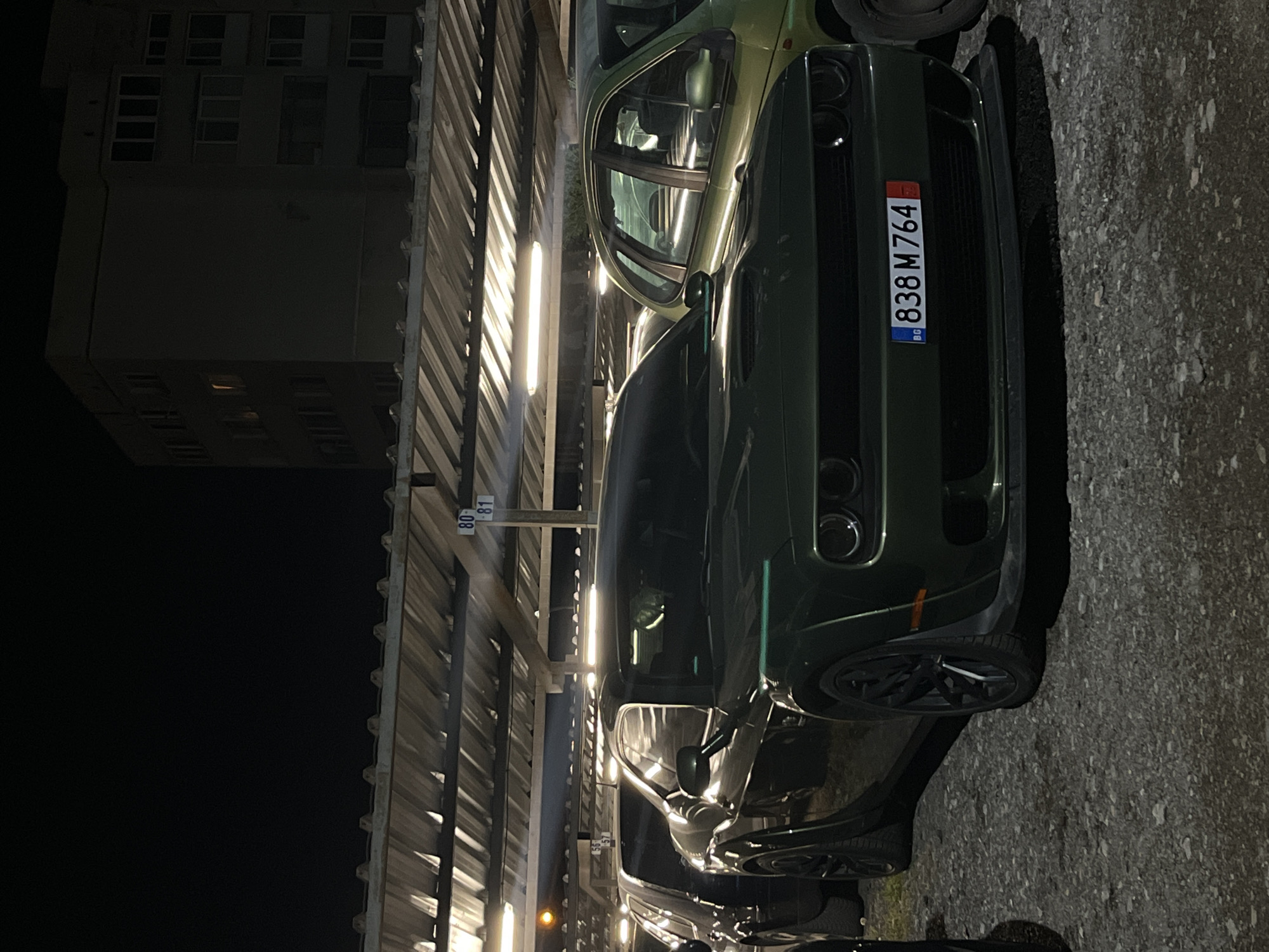 The image depicts a dark, nighttime scene, most likely an outdoor parking lot or covered parking area. A building can be seen in the background, but its details are obscured by darkness. The main subject of the image is a green car, possibly a Dodge Challenger, parked under a covered structure. The car's license plate reads "838 M 764" and is Bulgarian, indicating the location is in Bulgaria. The lighting is artificial, casting shadows from the parking structure. There is no visible human presence in the image. The environment appears to be urban. Based on the license plate, it is likely taken in Sofia, Bulgaria.