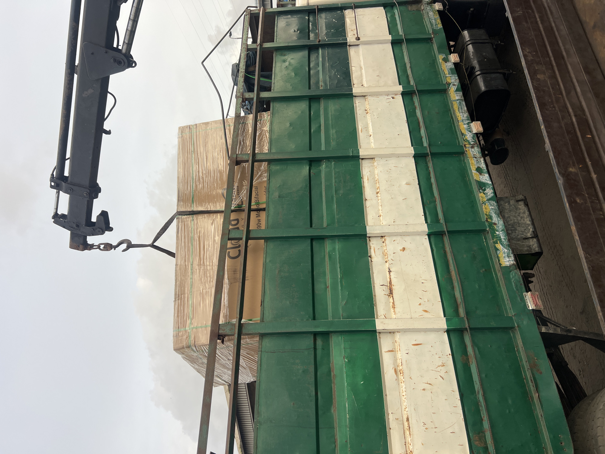 The image shows a truck with green and white painted panels, reminiscent of the Nigerian flag, loaded with wrapped rectangular packages. A crane with a hook is positioned to lift the load. The setting appears to be a construction or industrial area, possibly in Lagos, Nigeria given the color of the truck and the content of the packages, based on prior knowledge of shipping patterns. No people are visible. The sky is overcast. The focus is on the loading process.