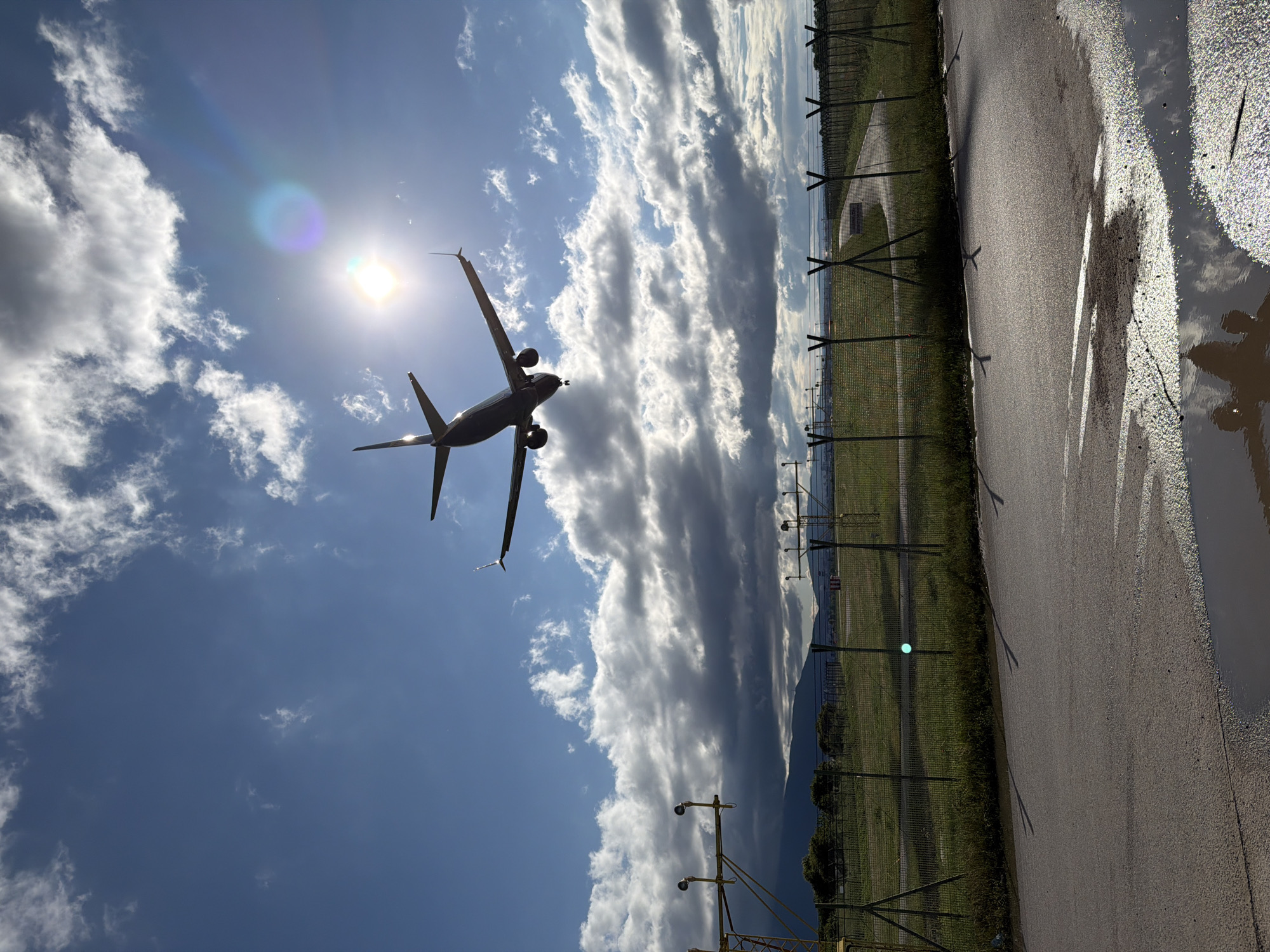 An airplane is captured in mid-flight, silhouetted against a bright, partly cloudy sky. The aircraft, a twin-engine jet, is seen from below, ascending or descending with its nose slightly pitched upwards, wings extending towards the edges of the frame. The sun, a brilliant white orb with a prominent lens flare and rainbow-colored halo, is positioned near the plane's nose, casting the aircraft in stark silhouette.
The sky is a vibrant blue, featuring scattered white cumulus clouds on the left and large, textured, and somewhat darker cloud formations stretching across the upper right.
In the foreground, a stretch of asphalt or tarmac occupies the lower portion of the image. On the right side of the tarmac, several large puddles reflect the bright sky and clouds above, suggesting recent rain. A tall chain-link fence, topped with barbed wire, runs horizontally across the middle ground, separating the paved area from a lush green field beyond. Fence posts cast long, distinct shadows across the tarmac, indicating a high sun angle.
Beyond the fence, various airport lighting fixtures and poles are visible in the grassy expanse. In the far distance, a range of mountains rises subtly against the horizon, partially veiled by atmospheric haze. There is no visible text. The scene depicts an airport environment in Sofia, Bulgaria, likely during midday, under dynamic weather conditions.