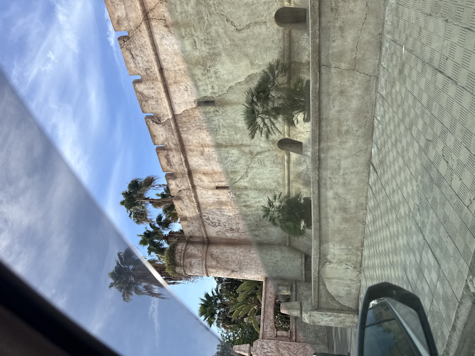An architectural facade with faux battlements extends into a blue sky dotted with cirrus clouds. Palm trees peek over the wall. The structure features textured, aged stone and decorative corbels. The photograph, possibly taken from a moving vehicle, captures a reflection in the car window, obscuring the bottom portion of the scene. Paved ground is visible on the right.