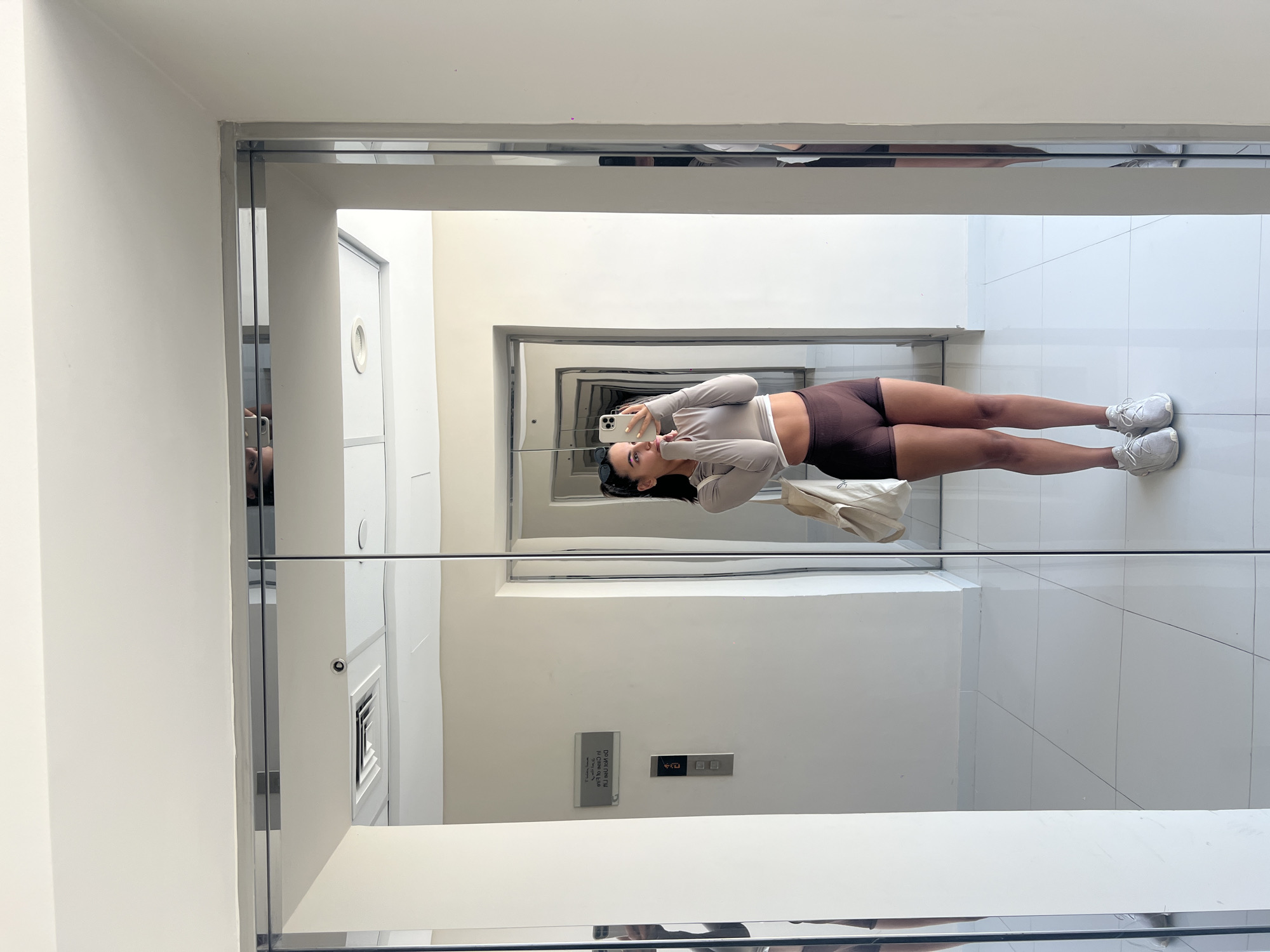A vertical full shot depicts a woman taking a mirror selfie in what appears to be an elevator in Dubai, United Arab Emirates. The woman is standing in the elevator wearing a long-sleeved light grey crop top and brown athletic shorts. She is holding a white smartphone and looking at the camera, with her right hand on her cheek. She is also wearing white sneakers. A beige tote bag hangs off her right shoulder.
The elevator walls are white, and the floors are tiled. A small metal sign and an elevator call button panel are visible on the wall behind her. There is a long narrow hallway reflected in the mirror, which creates a perspective effect. The lighting is bright, giving a clean, modern aesthetic. An air vent is visible as well. The mirror’s edges have a chrome or steel finish.