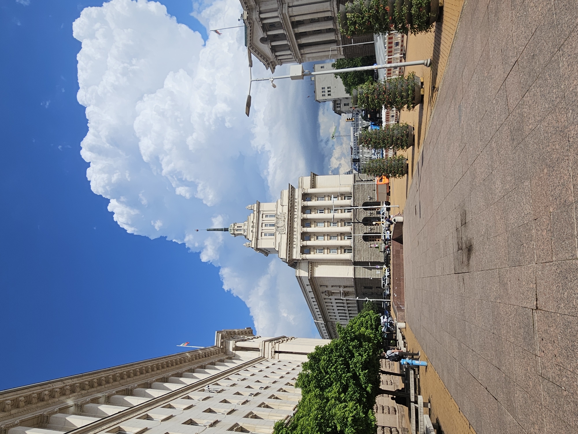 The image, rotated clockwise, depicts a wide urban street or square flanked by imposing classical-style buildings under a bright blue sky dotted with large, fluffy white cumulus clouds. The setting appears to be a sunny day.
In the center-right, a prominent, multi-story building features a tall spire and classical architectural elements, including columns and many windows. Directly to its left, another grand building with a similar architectural style, columns, and intricate detailing is visible. The text "НАРОДНО СЪБРАНИЕ" (Narodno Sabranie, meaning National Assembly) is clearly visible in Cyrillic script on this central building, indicating the location is the Parliament building in Sofia, Bulgaria. Further right, a third large building with a flat roofline and distinct window patterns can be seen.
The foreground features a wide, reddish-brown granite or stone paved area with visible seams, possibly a pedestrian zone or sidewalk. Along its edge, a lighter, sand-colored strip runs parallel to the buildings. Several small, green trees, some planted in decorative cylindrical planters, line the paved area.
A few individuals are scattered throughout the scene, primarily in the mid-ground, either walking or standing, suggesting typical daily activity in a public space. Other objects include streetlights, a lamppost, what appears to be a blue public utility box near the pavement edge, and parked vehicles visible further in the background. A red, white, and green flag (colors of the Bulgarian flag) can be seen flying from the top of one of the buildings. In the distance, a construction crane is faintly visible, hinting at ongoing development in the city.