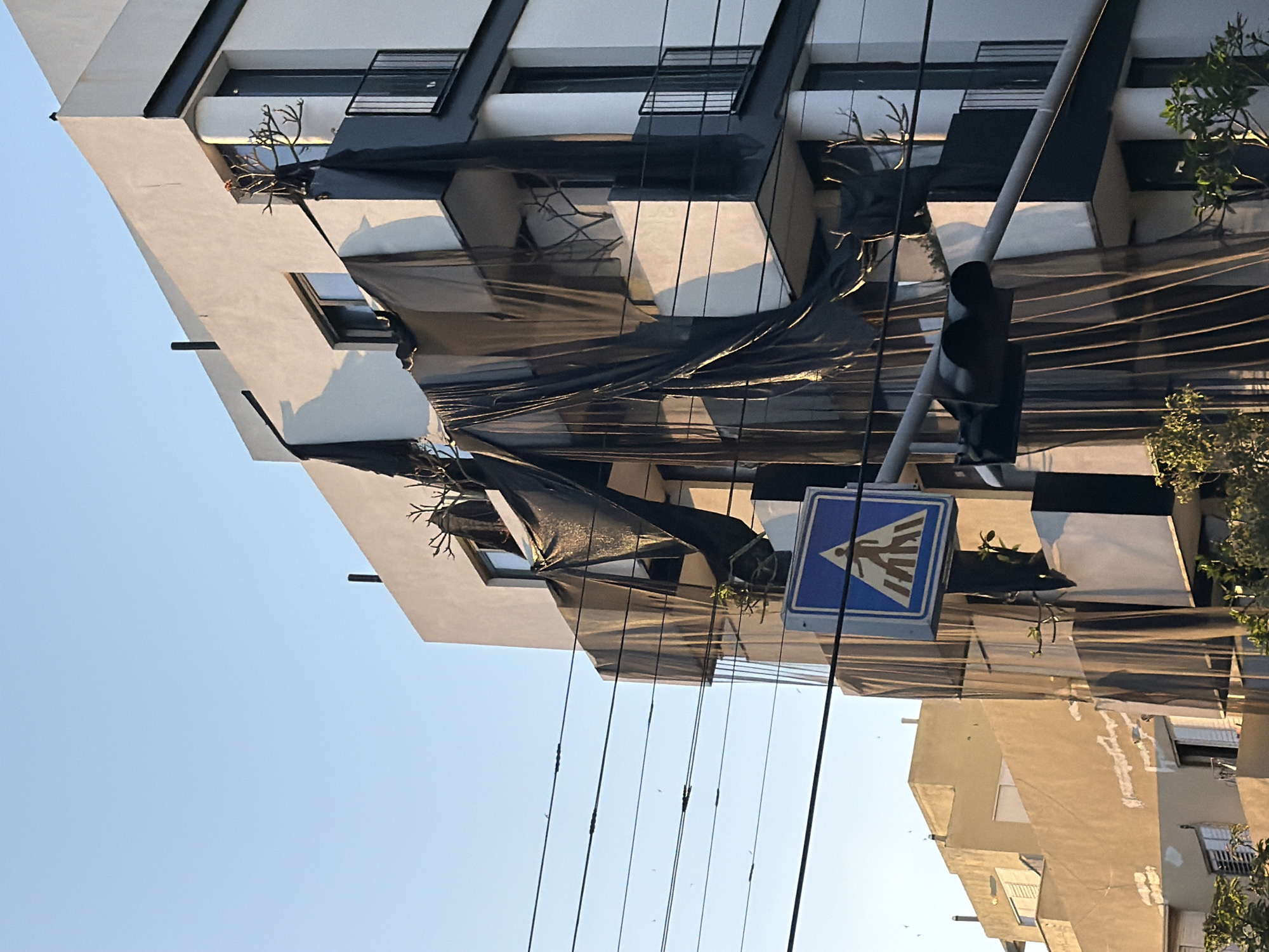A vertical photograph captures a building in Tel-Aviv, Israel, partially obscured by black mesh or netting draped over its angular balconies. A crosswalk sign stands in the foreground, partially blocking a clear view of the building behind it. Below, a low-rise building of a different design is visible in the background, along with a set of electrical wires. The sky is pale blue.