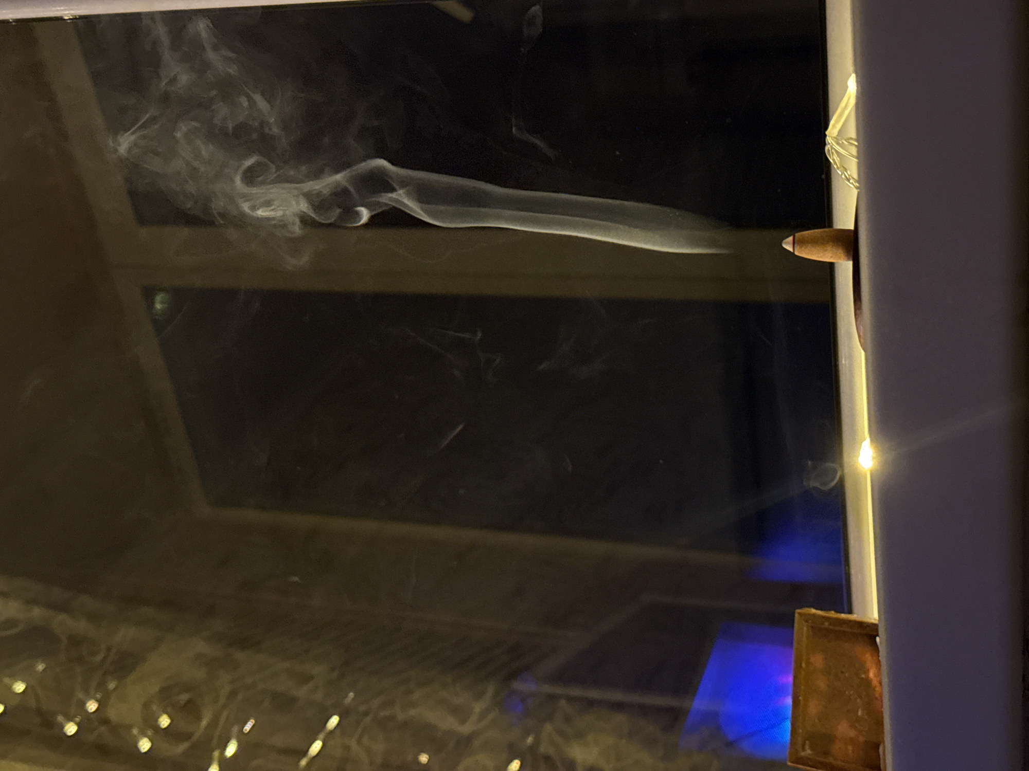 The scene depicts an interior view looking out a window at night in Naberezhnye Chelny, Russia. In the foreground, a light-colored window sill extends horizontally across the lower portion of the frame. On this sill, from left to right, a small, rectangular, tarnished metallic object (possibly copper) rests. To its right, a dark circular base supports an inverted conical incense burner, which is light brown with a dark burning tip and a narrow white band. A distinct column of white-to-light-grey smoke rises vertically from the burning incense, then curls and dissipates into fluid, swirling patterns toward the upper right of the frame. Further to the right on the sill, a small, clear, irregularly shaped object, possibly coiled wire or a piece of plastic, is visible. The window glass, acting as a reflective surface in the mid-ground, shows prominent reflections of the interior. These reflections include a vertical string of unfocused, warm-toned lights on the left side, and beneath them, a rectangular blue light source. Through the window, the exterior is predominantly dark, indicating nighttime, with faint, indistinguishable outlines of what appears to be part of a building's structure or an external window frame visible in the distance. No individuals are present in the image.