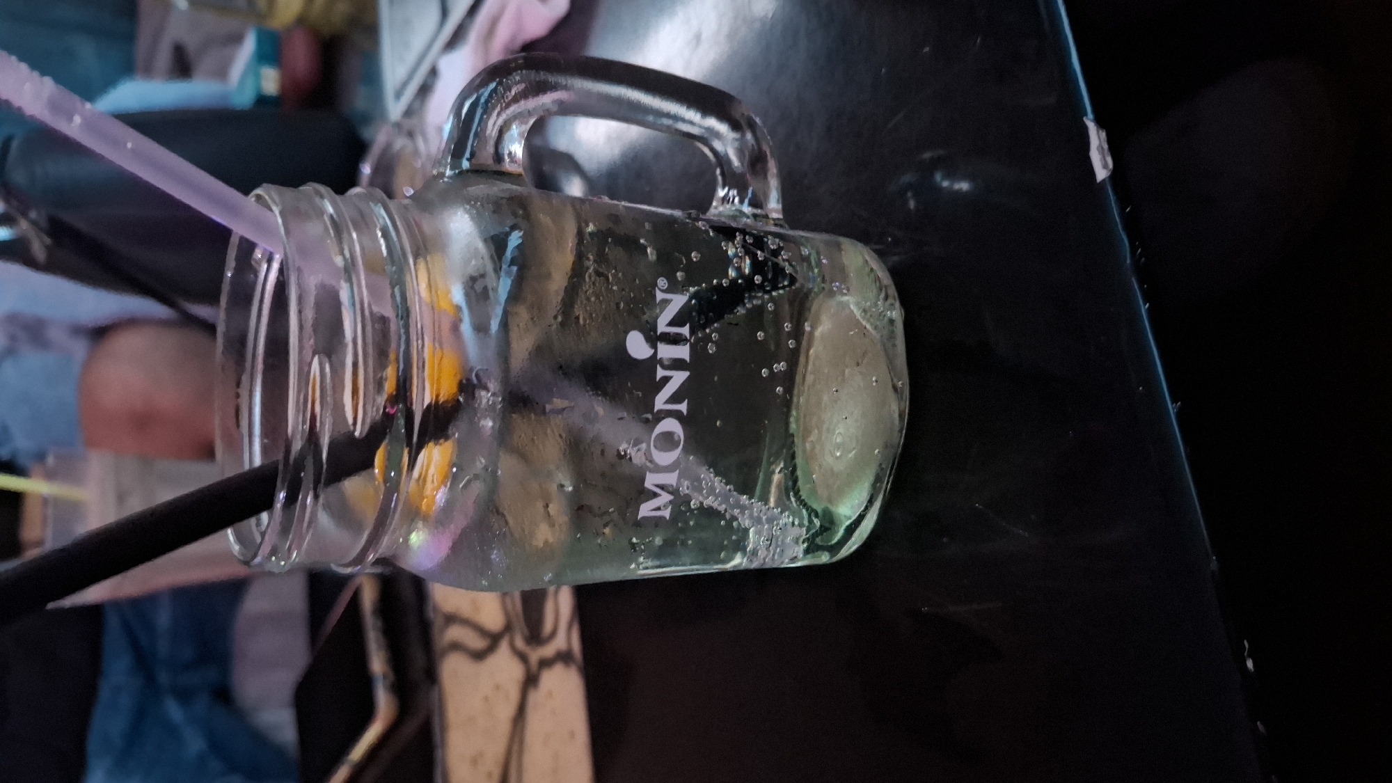 A glass mason jar-style mug filled with a clear liquid and adorned with the "MONIN" brand logo is positioned on a dark, reflective surface, likely a table. The mug has a metal handle and two straws inserted into it: one black and one translucent pink. Inside the mug, there appear to be pieces of fruit, possibly citrus. The background is blurry, but suggests an interior setting with glimpses of other objects and surfaces. This image does not definitively provide information that would indicate the location is in Svishtov, Bulgaria. The image shows a refreshment drink on a table, indoors.