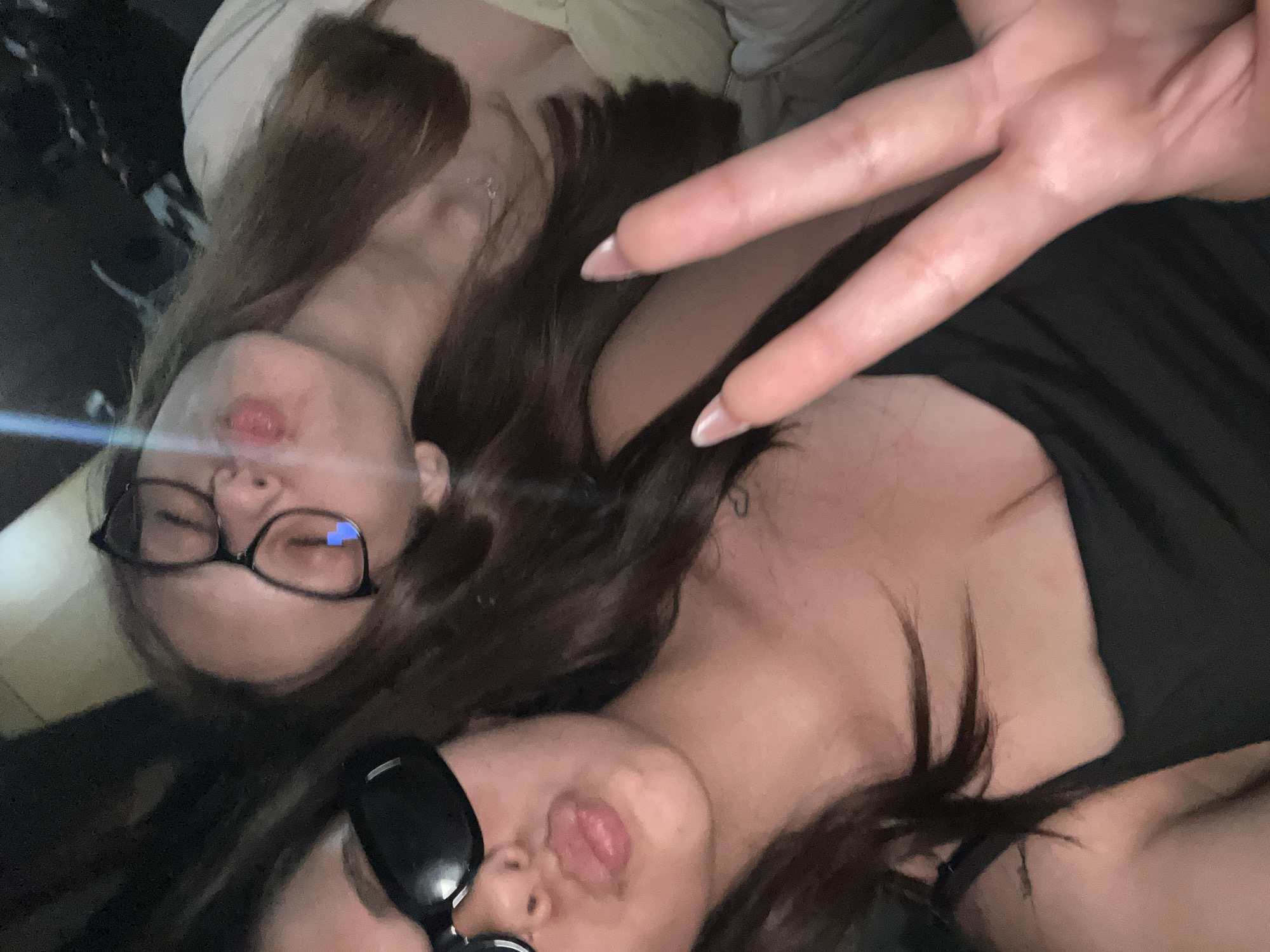 Two young adult females are depicted in a close-up, angled selfie. The scene is dimly lit with artificial light sources creating lens flares.
The female in the foreground, positioned on the left, has dark, wavy hair. She wears black sunglasses and a black spaghetti strap top. Her lips are slightly parted, and her tongue protrudes to the left. A small, faint heart-shaped tattoo is visible on her upper right chest/shoulder area. Her left hand is partially visible, holding what appears to be the camera device.
The second female, positioned in the background to the right, has long, straight brown hair and wears black-framed prescription glasses. A digital blue heart emoji is overlaid on the left lens of her glasses. Her lips are pursed, forming a "fish face" or blowing a kiss. She wears a light-colored, possibly cream or white, top. Her right hand is extended into the lower foreground, forming a two-finger peace sign. Her fingers exhibit long, almond-shaped nails with a natural pinkish-nude color, indicative of artificial nail enhancements.
The background is dark and indistinct, suggesting an interior space or vehicle cabin at night. A lighter, out-of-focus surface is visible in the upper background. Location is Unknown, Unknown.