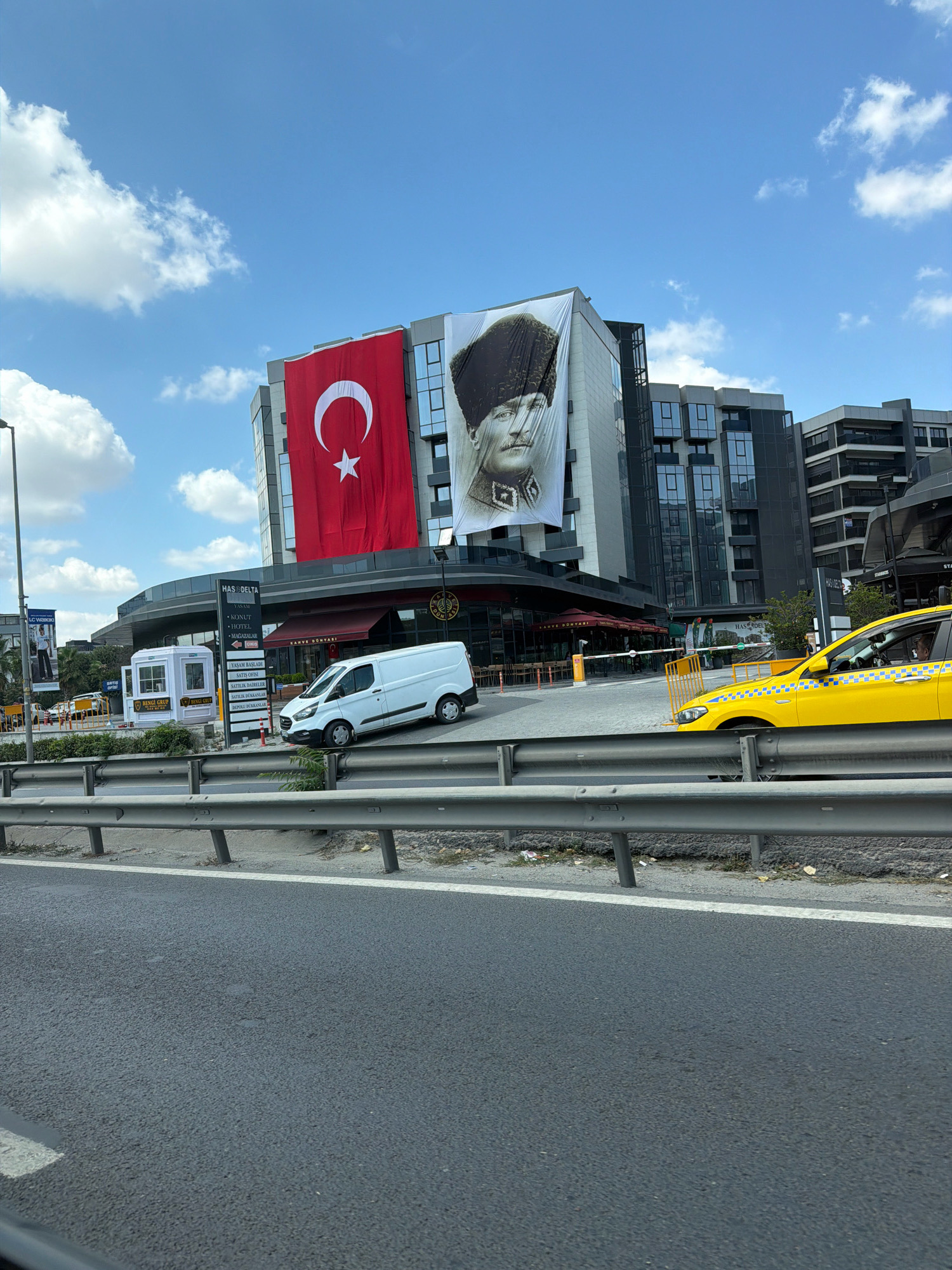 The image depicts a wide, slightly tilted shot of a street scene in Bahçelievler, Turkey, dominated by a large building featuring a prominent Turkish flag and a portrait of Mustafa Kemal Atatürk. The building, modern in design with multiple stories and glass windows, displays a vibrant red flag with a white star and crescent. To its right, a black and white portrait of Atatürk, wearing a traditional hat, is also displayed on the building's facade.
In front of the building, a white commercial van is parked on the street, positioned at an angle. To the right of the van, a yellow taxi is visible, its front end facing the viewer. The street is lined with a metal guardrail, and the foreground shows a section of asphalt road. The sky above is a bright blue with scattered white clouds.
Several signs are affixed to the building. One sign, partially obscured, reads "HAS.DELTA" and lists various services including "VAN", "KONUT", "HOTEL", and "MAĞAZALAR." Another sign indicates "KAHVE DÜNYASI" (Coffee World).
The overall impression is of a busy urban environment in Turkey, marked by patriotic symbols and commercial activity. The presence of the van and taxi suggests ongoing traffic and daily operations. The building itself appears to be a mixed-use commercial or residential complex.