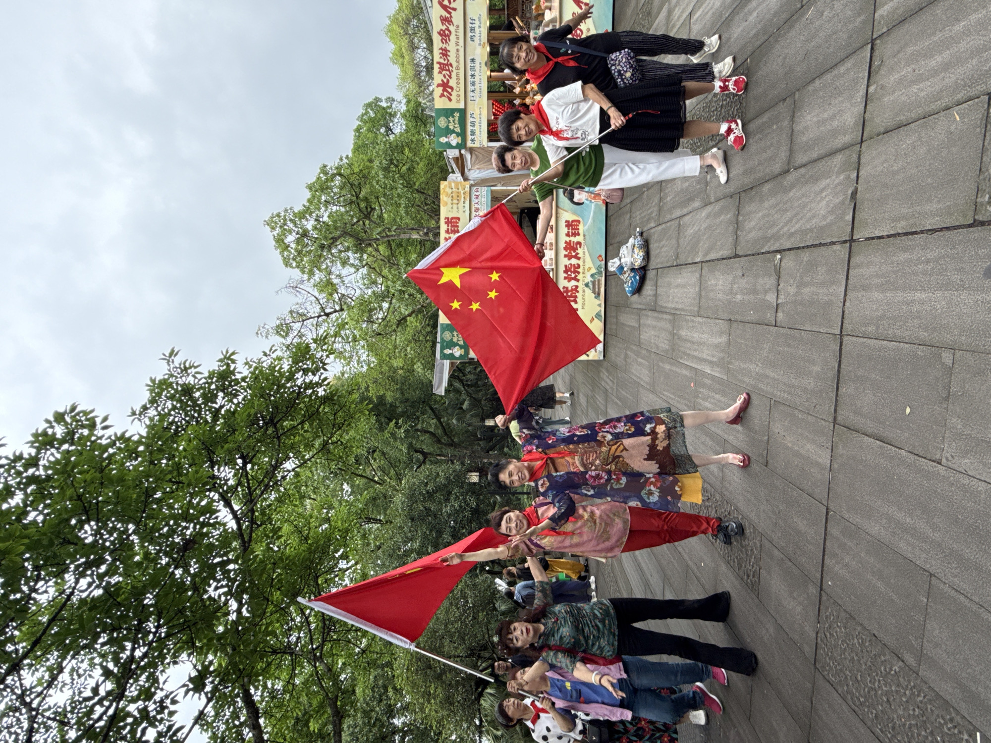 The image depicts a group of elderly women posing with Chinese flags in what appears to be a public space. Seven women are lined up on a stone-paved area, each holding a red flag with yellow stars, the national flag of China. The flags are slightly wrinkled as if they've been handled.
The women are dressed in a variety of casual clothing, suggesting they may be on an outing or engaging in a public activity. Some of them are wearing red scarves, which may indicate a connection to a particular group or event. They are smiling and looking towards the camera. Behind them, there are food stalls with signs in Chinese characters, indicating that they are in a public market or tourist area. Based on the signage, the location is Yuzhong District, China. There is also a lot of greenery, with trees in the background, giving the impression that the photo was taken on a cloudy day.