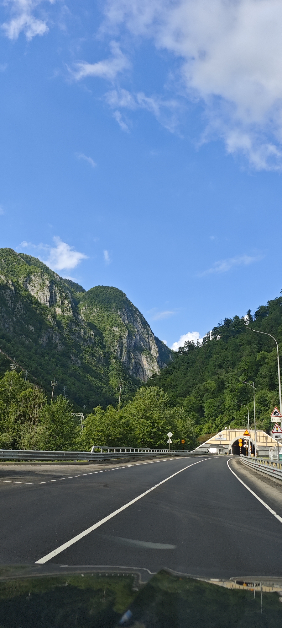 A vertical, eye-level shot depicts a two-lane road leading to a tunnel entrance in the Monastyr district of Russia. The road is marked with a solid white center line. A metal guardrail runs along the left side of the road, with dense green trees and vegetation behind it. Towering mountains rise in the background, partially covered in lush greenery and showing rocky cliffs. The sky is a clear blue with scattered white clouds. The tunnel entrance is cream-colored with ornate decorations above it, and at least three individuals stand near the entrance. Several road signs are visible to the right of the road, near the tunnel. The bottom of the shot reveals a small portion of the car's dashboard from where the photo was taken.