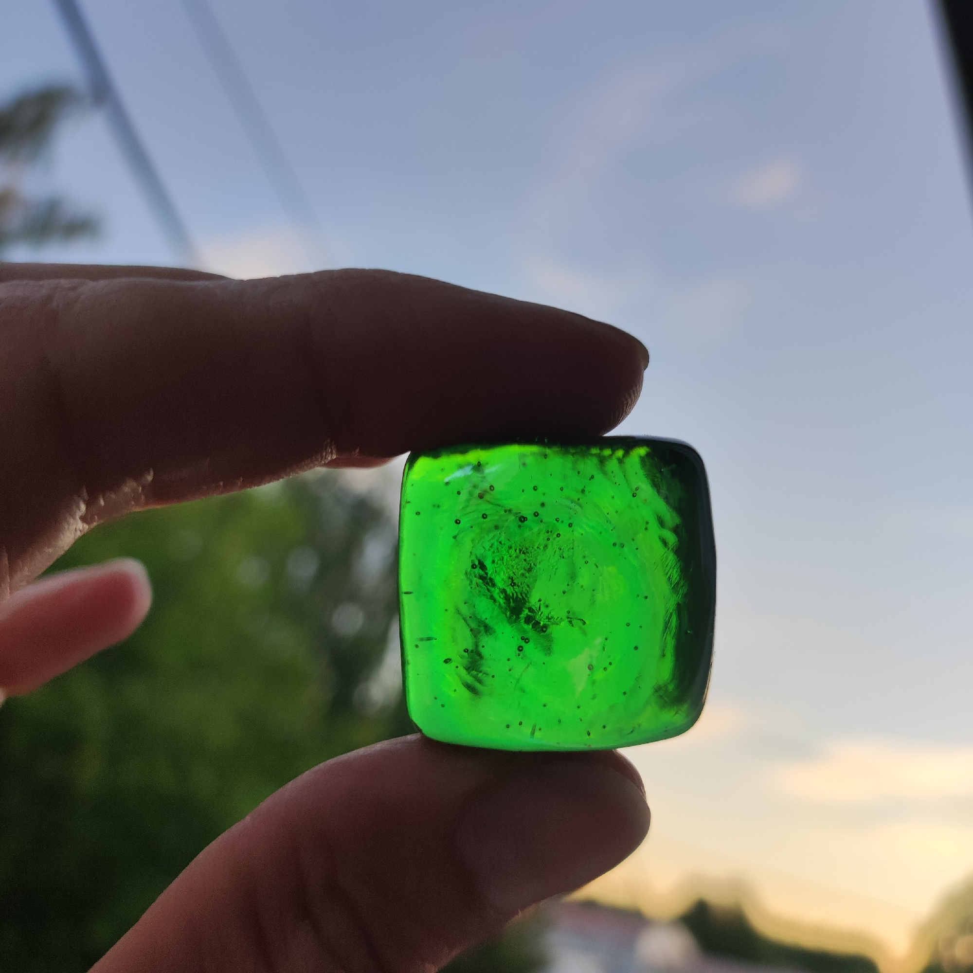 A close-up, horizontal, eye-level shot shows a person's fingers holding a translucent green, square-shaped object against a blurred background of sky, trees, and buildings. The object has small bubbles and imperfections within it. The sky is a pale blue with some wispy white clouds, and the sun appears to be setting or rising, casting a warm glow. The overall scene is softly focused, with the sharpest detail on the object and the fingers holding it. The location is Kineshma, Russia.