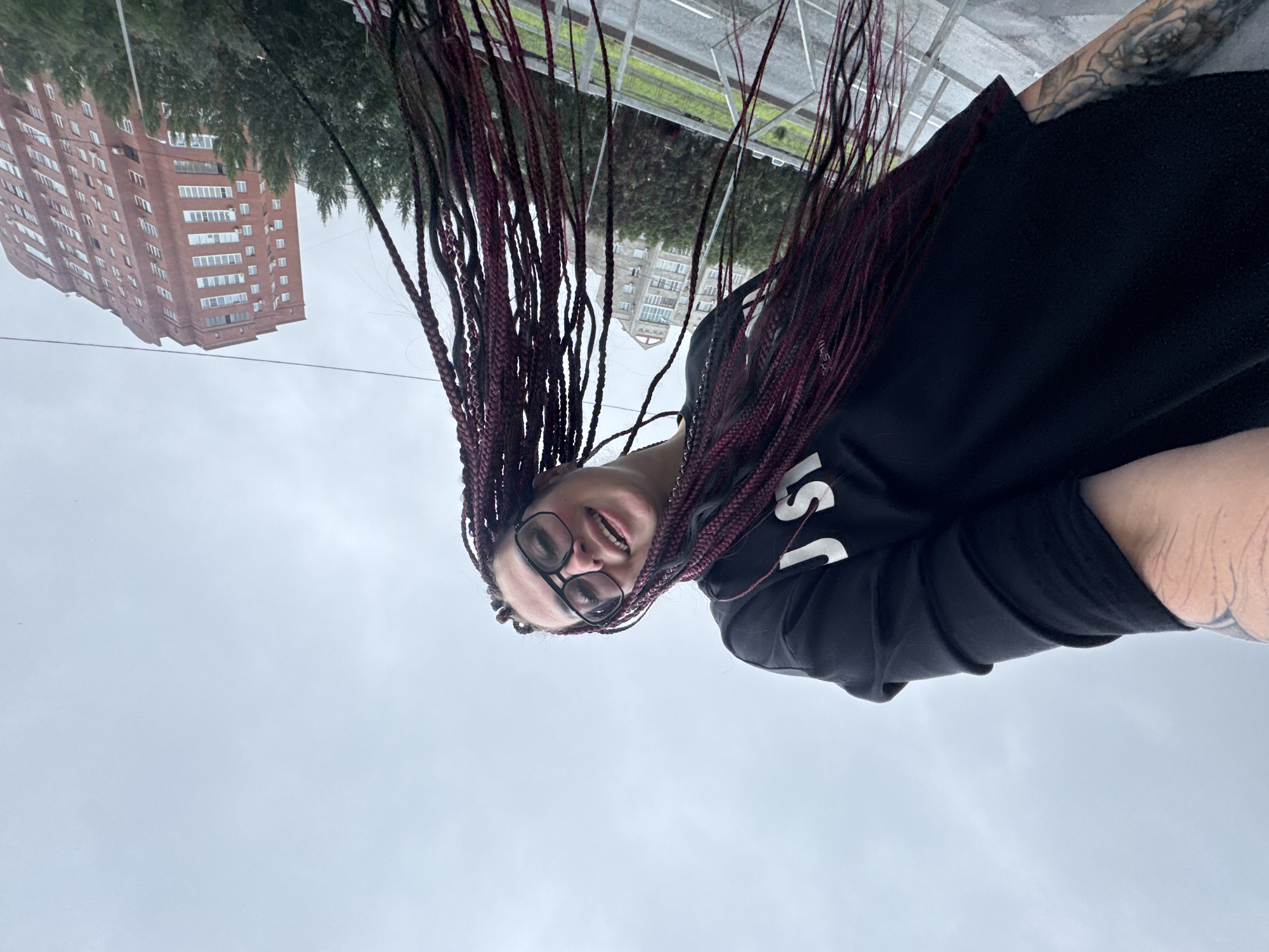 The image shows a young woman with braided, dark red hair and glasses, wearing a black sweatshirt. She is smiling and looking up, with her hair blowing in the wind. In the background, a tall, red brick building is visible, along with trees and a cloudy sky. The scene appears to be an outdoor location, possibly in the city of Tolyatti, Russia, given the architectural style of the building. The woman has tattoos on her arm.
