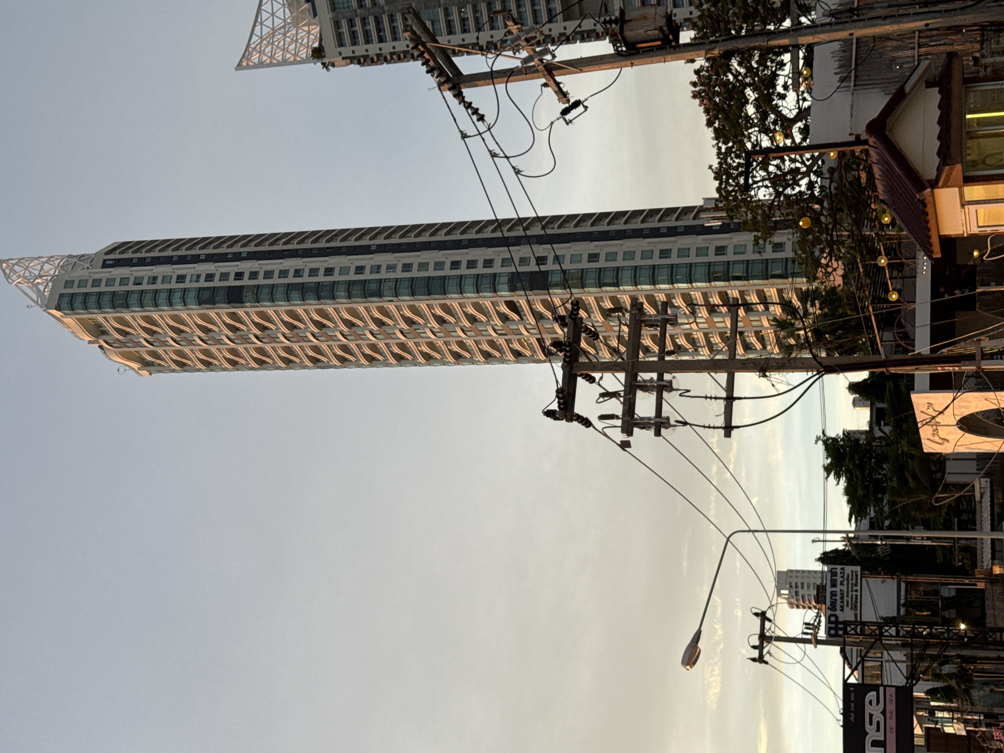 Captured from a low angle in Pattaya City, Thailand, the image showcases a tall, modern building with a distinctive, patterned facade reaching into a pale sky. Dense clusters of power lines and utility poles dominate the foreground, framing the architecture. A street lamp stands prominently to the side, while signage and partial views of nearby structures hint at commercial activity in the immediate vicinity. The lighting suggests either dusk or dawn.