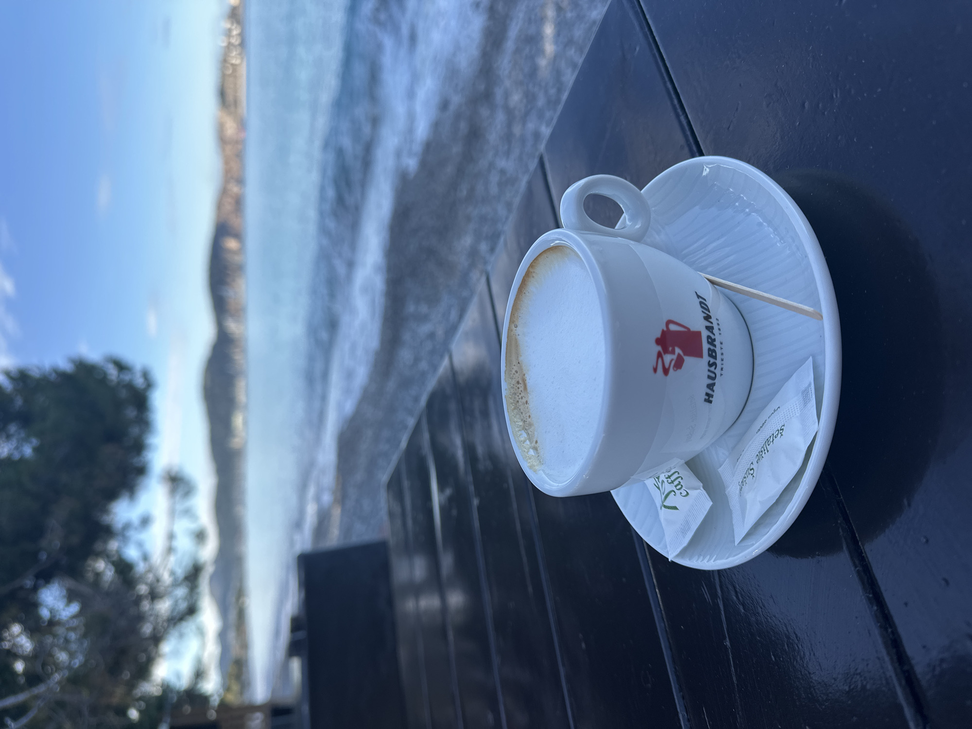 A cup of coffee with a white foamy top rests on a saucer with two sugar packets and a wooden stirrer. The cup, bearing the brand name HAUSBRANDT, sits on a dark, wooden surface, likely a table. In the background, a blurred view of the sea and a coastline is visible, along with a few trees. Based on the location, this could be a coffee served at a cafe along the beach in Bar, Montenegro.