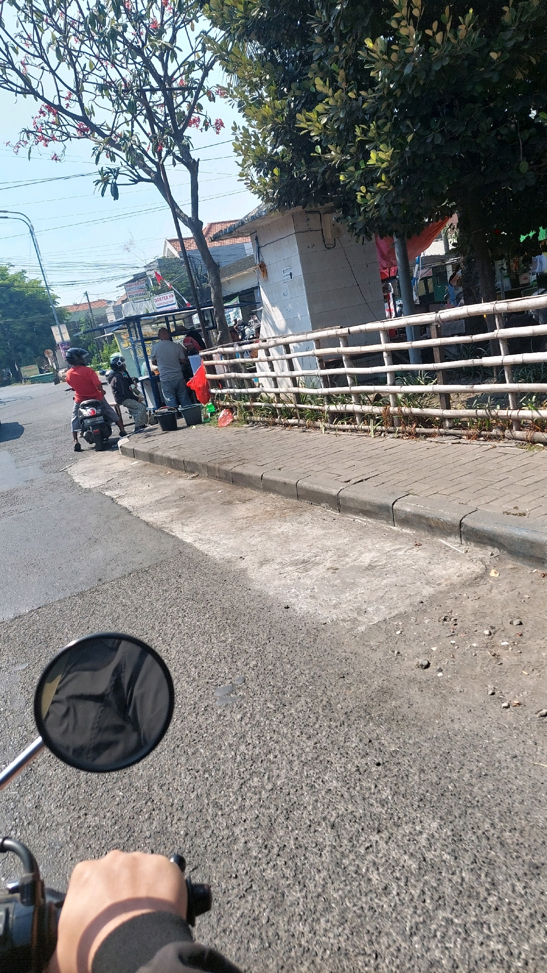 The image captures a scene on a street in Sepande, Indonesia, on a bright, sunny day. The perspective is from someone riding a motorcycle, indicated by the handlebar and rearview mirror in the foreground. The road surface is asphalt. To the right of the road, there is a sidewalk paved with gray bricks, bordered by a concrete curb. Beyond the sidewalk is a bamboo fence, and behind that, a small white building with a red awning. Trees with green foliage and some red flowers are visible, along with power lines and buildings in the background. On the sidewalk, near the bamboo fence, are two black buckets and a red plastic bag. A man in a gray t-shirt and jeans stands near the building. Two people are on a motorcycle parked on the side of the road. The person in front is wearing a red shirt and a helmet, and the person behind is wearing a black jacket and a helmet. The overall impression is that of a typical street scene in a tropical urban or semi-urban environment.