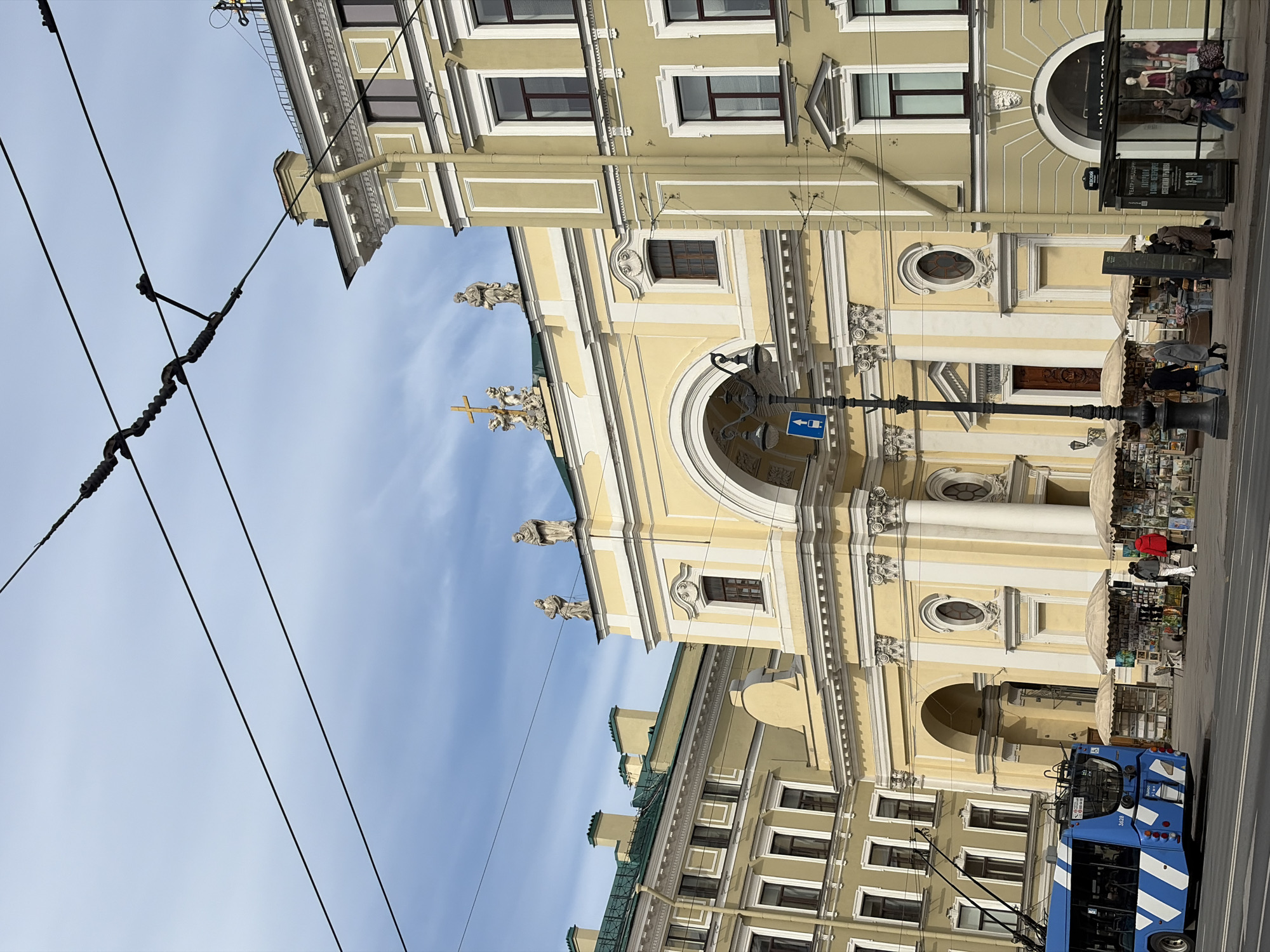 The image shows a street view in Saint Petersburg, Russia. The architecture features a large, ornate yellow building with white trim, a prominent archway, statues on top, and a cross. The building is bordered by other buildings with traditional window designs. Street vendors are selling goods in front of the building. People are browsing the vendor stalls and walking on the street. A blue and white trolley bus is visible on the right side of the frame. Overhead power lines are visible in the foreground against a blue sky with some clouds.