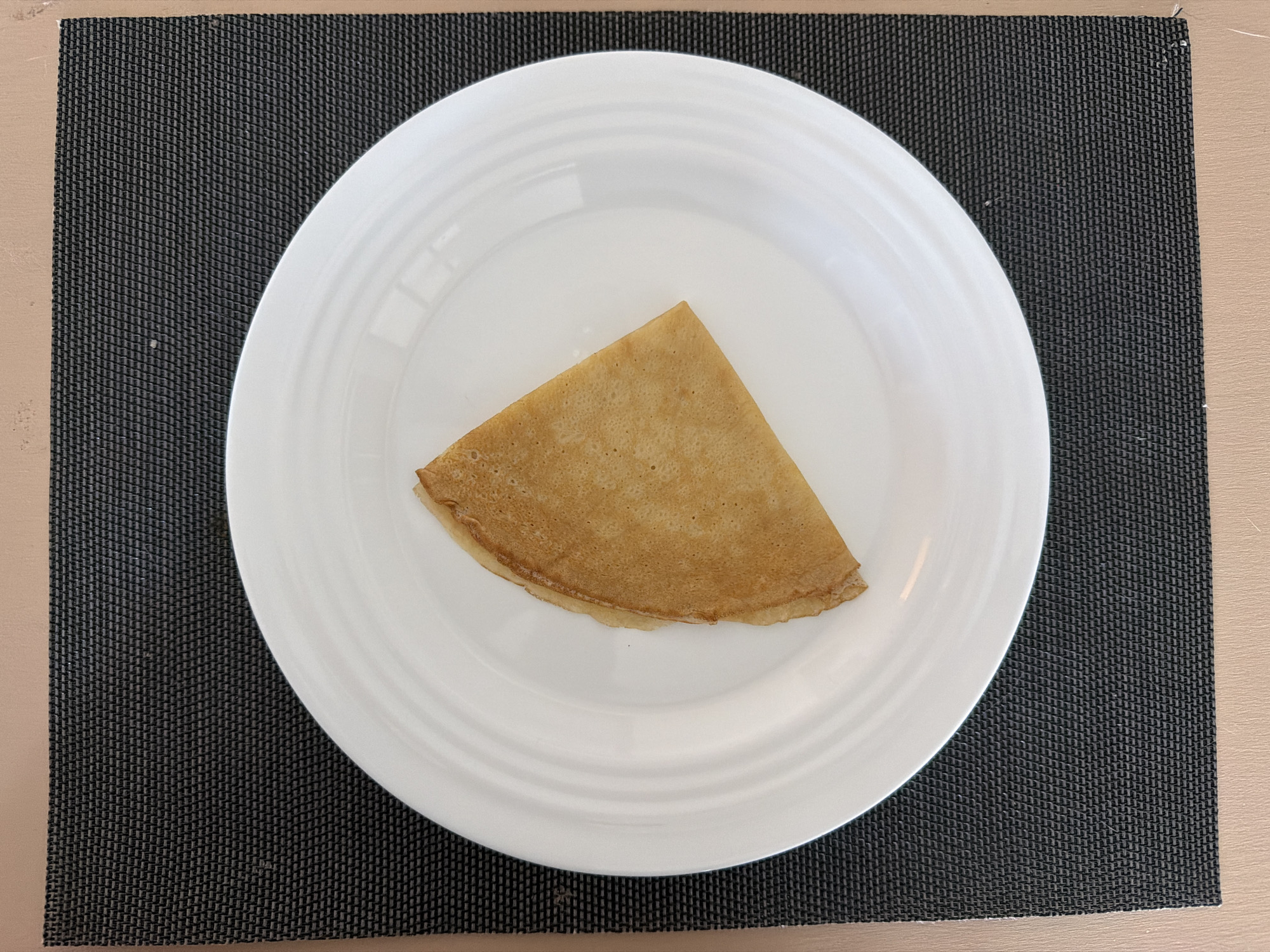 A single, folded crepe rests on a white plate, which is positioned on a dark gray placemat. The placemat is on a light brown surface. The image is a top-down view. There are no people or animals present. The background is plain and offers no discernible location information.