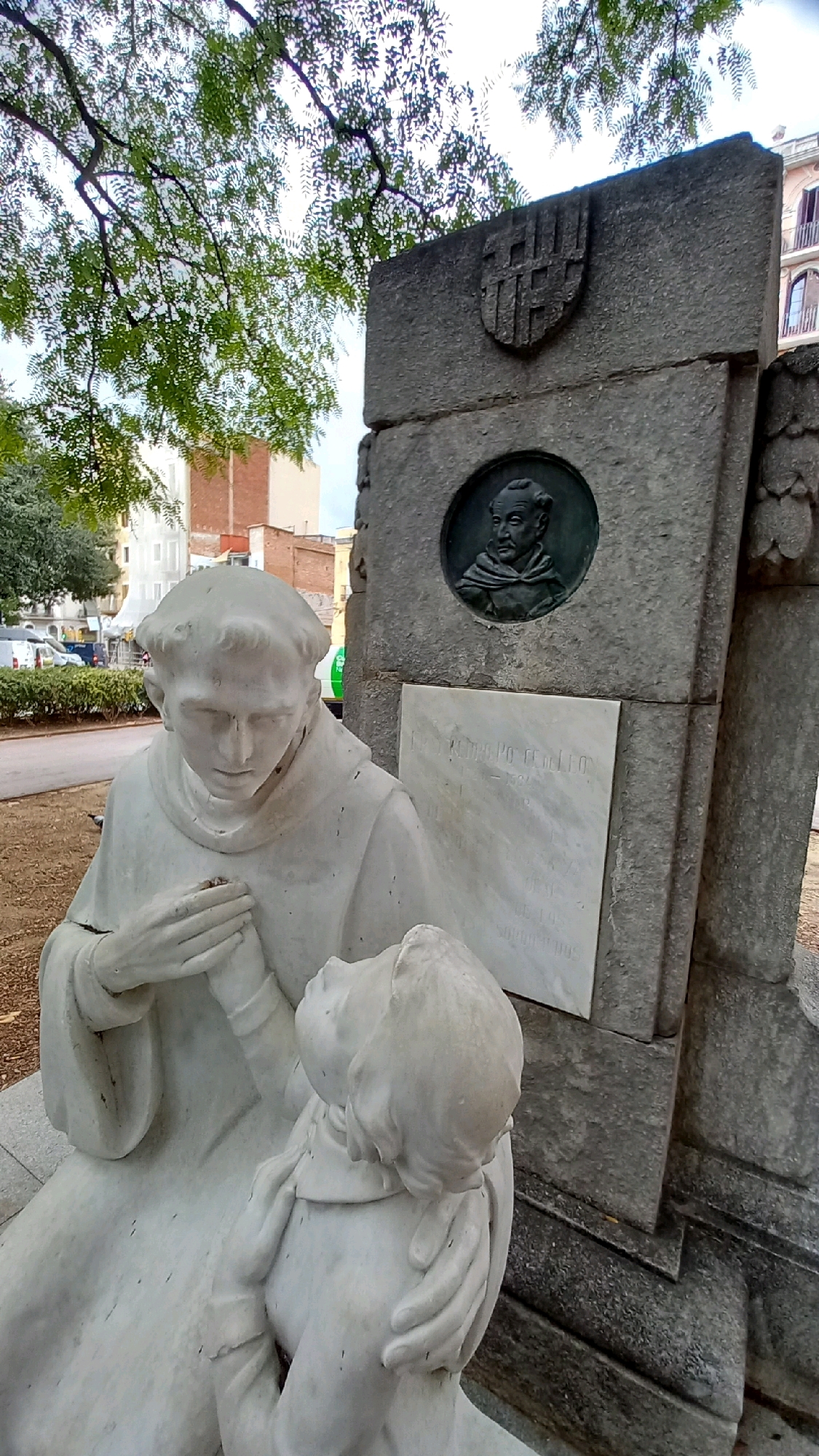 The image shows a sculpture of two figures in what appears to be a park or public square in Barcelona, Spain. The sculpture, made of a light-colored material, depicts a robed figure with hands clasped, looking down at a smaller figure kneeling in front of it. The larger figure appears to be a friar or monk. Behind the sculpture is a large stone monument with a carved crest and a circular bronze plaque with a bust of a man. A marble plaque with text is also visible on the monument. The background shows trees, buildings, and a street with parked vehicles, suggesting an urban setting. The monument is likely a memorial.