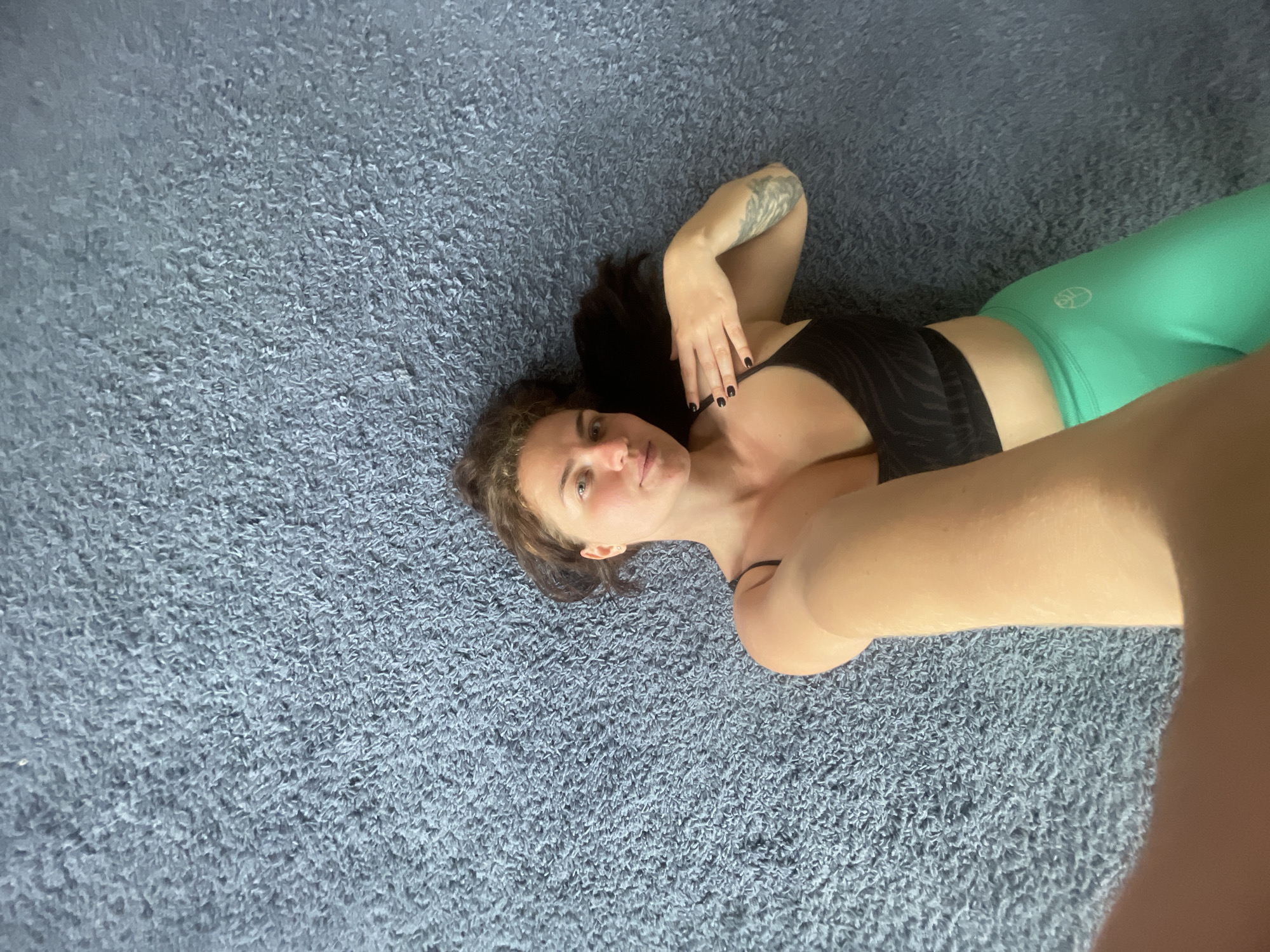 A woman is lying on a textured blue rug. She is wearing a black sports bra and bright green leggings. The woman has dark hair and a tattoo on her left arm. Her nails are painted black. She is looking at the camera, holding her right arm up, taking a selfie. The background is the blue rug, which fills most of the frame. The location cannot be determined from this image.