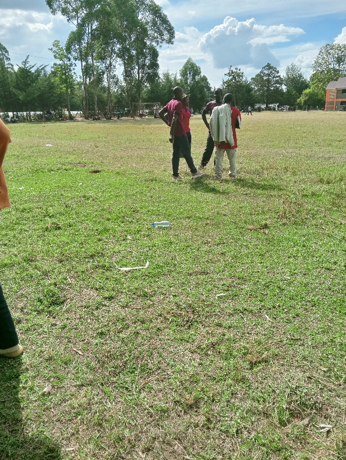 The image depicts an open, partially dry grassy field in Lwandeti ward, Kenya, under a blue sky with scattered white cumulus clouds. In the midground, three adult males are standing together. The male on the left wears a maroon short-sleeved shirt, dark trousers, and light-colored shoes, with his right hand placed on his hip. Next to him, a second male, also in a maroon short-sleeved shirt and dark trousers, faces towards the left. The third male, positioned to the right of the group with his back partially to the viewer, wears a light-colored long-sleeved jacket draped over his shoulders, over a red shirt, and light-colored trousers. The foreground shows predominantly green and dry grass, containing a clear plastic water bottle and fragments of white paper. A partial view of a person's leg and light-colored footwear is visible in the lower-left corner. The background features a large expanse of similar grassy terrain, bordered by a dense line of trees, including tall eucalyptus trees, on the left. A multi-story orange building is visible in the far background on the right. Distant, indistinct figures are present across the field, and a metal structure resembling a goalpost is discernible near the distant treeline on the left.