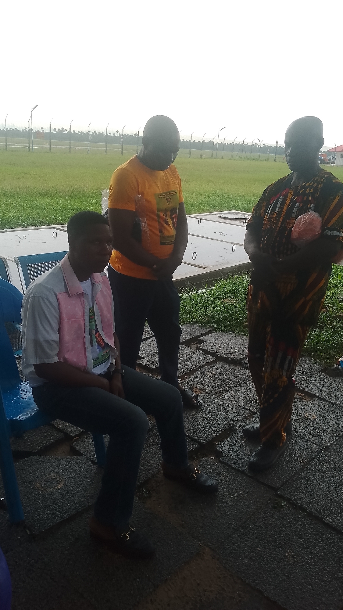 Three individuals are present in an outdoor, overcast environment, which appears to be an airfield or similar open area, possibly in Asubi, Nigeria.
On the left, a young man is seated on a blue plastic chair. He is wearing a light-colored collared shirt with pink detailing on the front and a white t-shirt underneath. He has dark pants and black loafers with a gold buckle. He is looking to his right.
In the center, a man wearing an orange t-shirt with a graphic design and dark pants stands facing to his left. He has his hands clasped in front of him.
On the right, a man stands facing to his left, wearing a colorful, patterned traditional garment and dark shoes. He is holding a plastic bag in his right hand.
The ground is paved with dark interlocking stones, with some patches of wetness and vegetation. In the background, there is a wide expanse of green grass, a fence line, and distant trees. The sky is bright and overcast, indicating diffused light. Structures that appear to be manhole covers or utility access points are visible on the paved area.