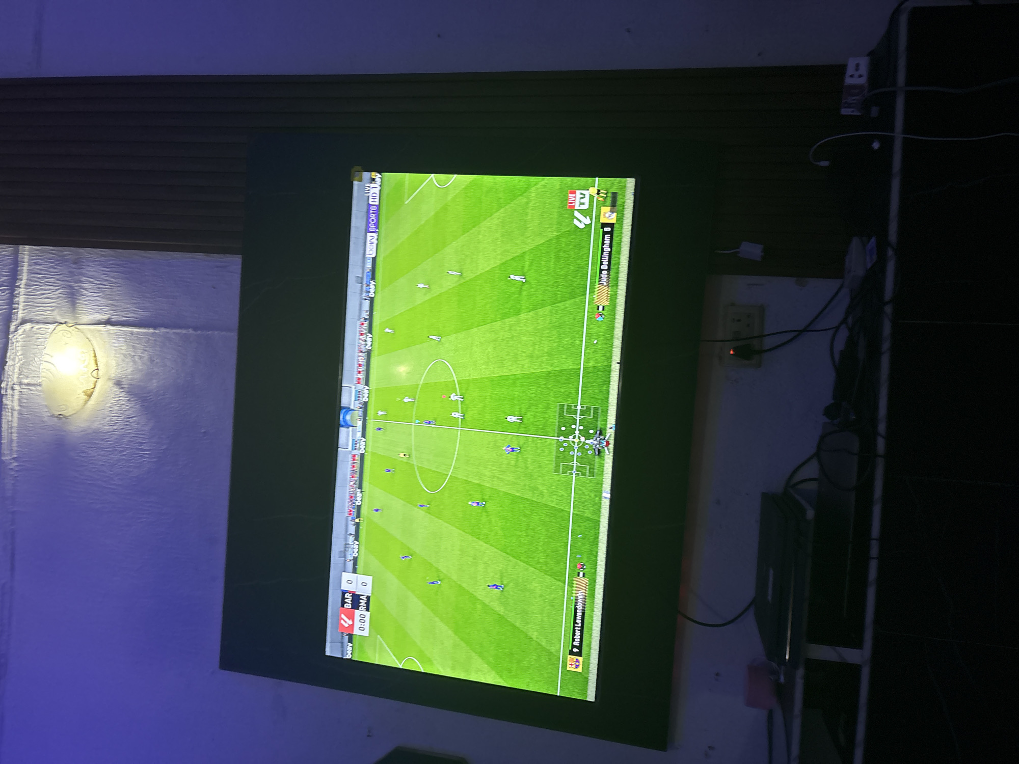 The image displays a television screen showing a football (soccer) match being played on a vibrant green field. The perspective is slightly angled, with the screen oriented vertically. The scoreboard at the bottom left indicates the teams are "BAR" and "RMA", both with a score of 0, and the time is 0:00. Text overlays on the screen show "beIN SPORTS HD 1" and "LIVE TV". Another overlay in the top right shows "Jude Bellingham B". At the bottom right, a player is identified as "Robert Lewandowski". The scene is set indoors, with the television mounted on a dark wall that transitions to a striped wooden paneling above. To the left of the screen, a white textured wall is visible with a lit light fixture. Electrical outlets and cables are present on the wall to the right of the television, suggesting a domestic or entertainment setting. The lighting appears to be dim, with a purple hue emanating from the left side of the frame, possibly from ambient room lighting. The overall impression is of someone watching a football game, likely a video game simulation given the graphical elements and scoreboard style. The location context is Obosi, Nigeria.