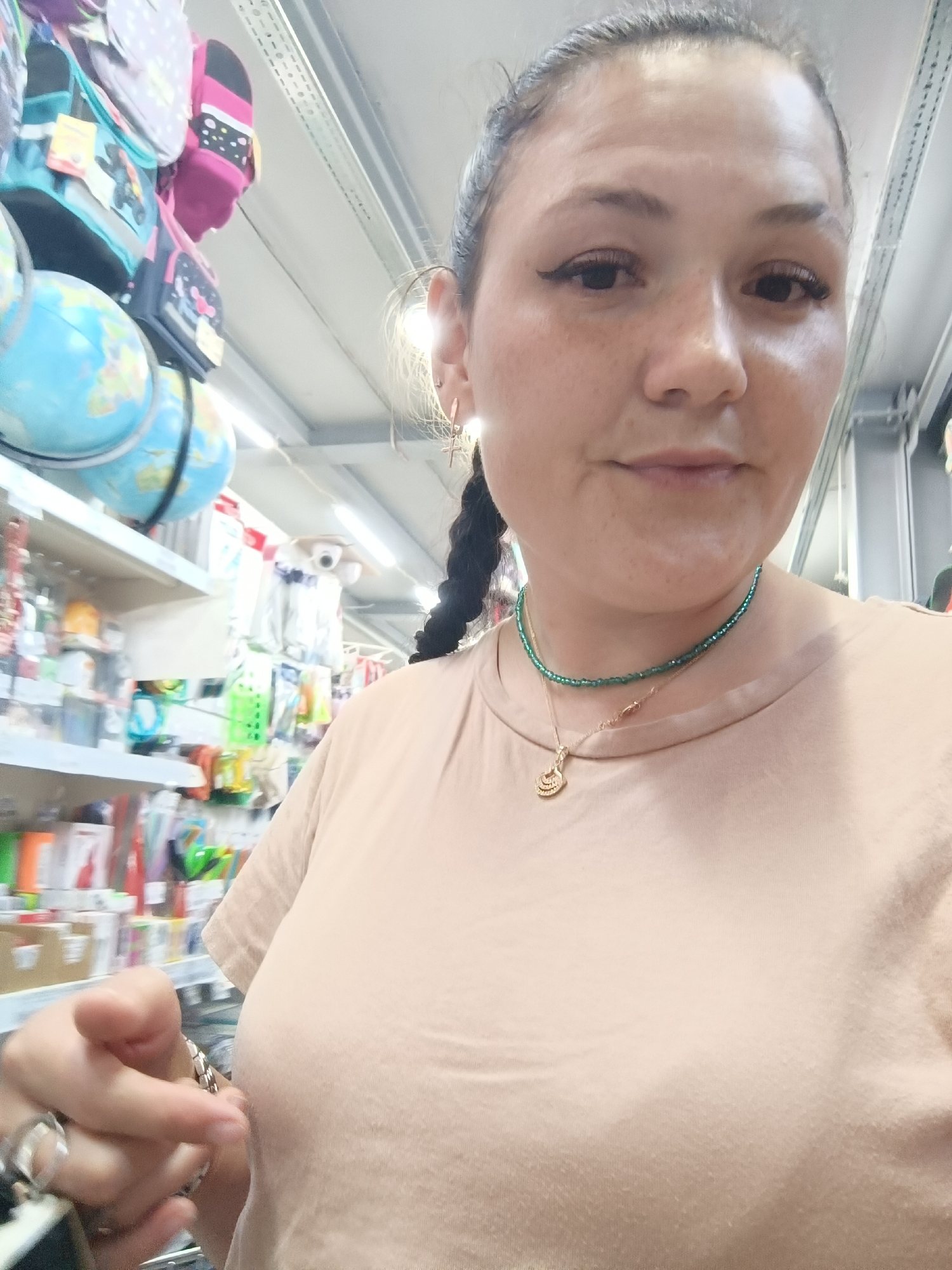 A woman stands in what appears to be a retail environment, possibly a store with shelves stocked with merchandise. The woman is the primary subject and is looking towards the camera, taking a selfie. She has dark hair styled in a braid and is wearing a light-colored t-shirt. Around her neck, she wears two necklaces, one green beaded and another with a gold pendant. Her left hand is extended towards the camera, and she is wearing several rings on her fingers. In the background, shelves are visible, displaying various items, including what appear to be globes and small backpacks. The lighting suggests an indoor setting, likely a well-lit retail space. The image does not provide specific information about the city of Chaikovsky, Russia, or any details that would indicate it is located there.