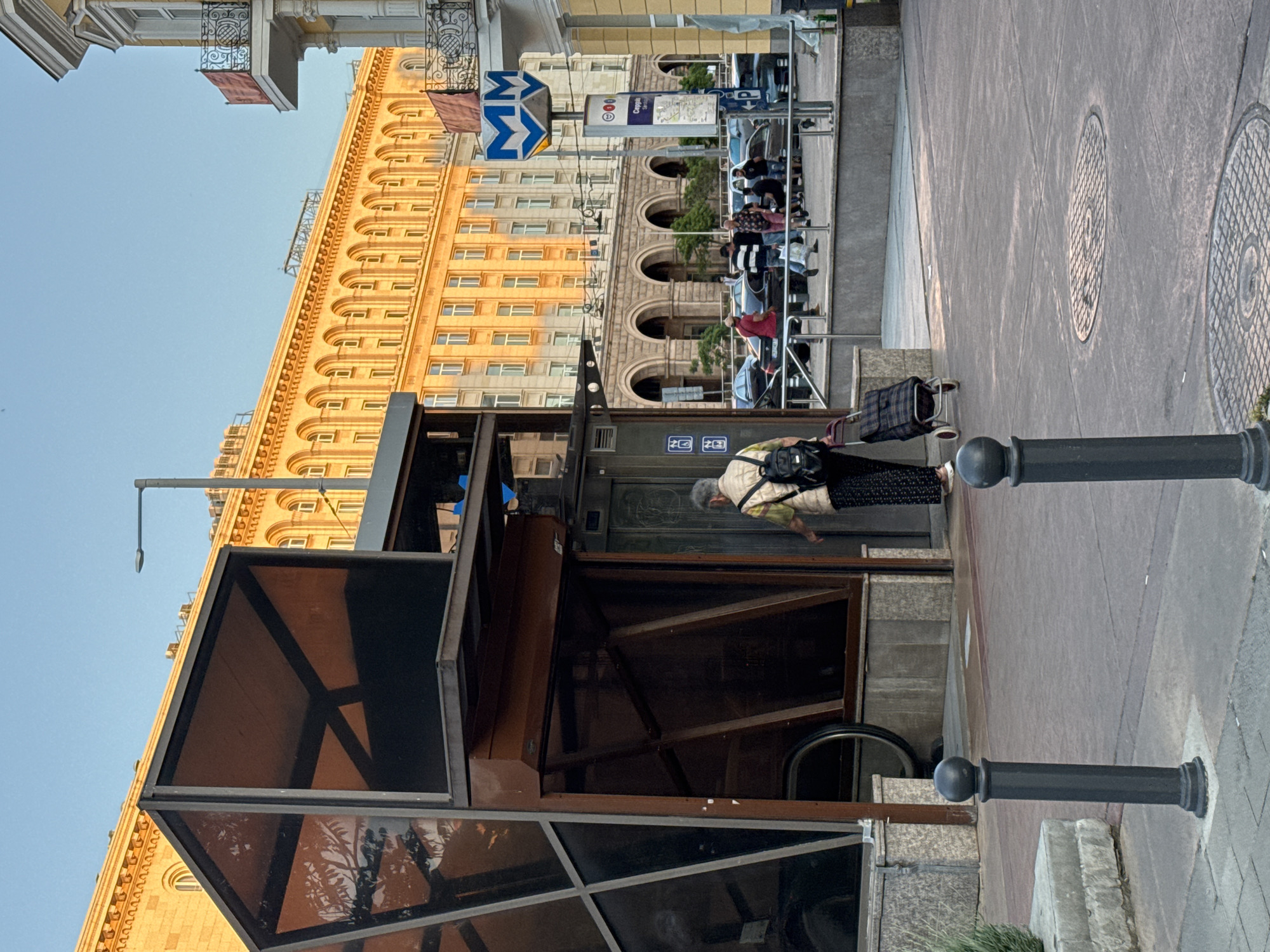 The image captures a street scene in Sofia, Bulgaria, featuring a woman walking towards the entrance of a metro station. The woman is pulling a rolling suitcase. The entrance is a modern glass and metal structure. In the background, a large, yellow-toned building with arched windows is visible, along with a metro sign. There are also dark metal posts along the sidewalk. The environment appears to be a bustling urban area.