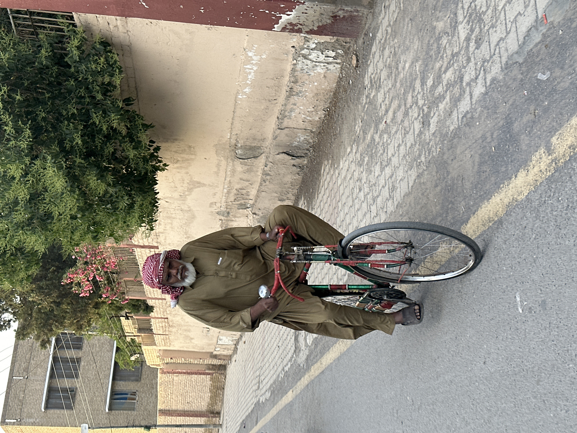 An elderly man is standing on a bicycle in a street in Quetta City Tehsil, Pakistan. He is wearing a traditional brown shalwar kameez and a red and white checkered keffiyeh. The bicycle is old and painted in red and green. The street is paved and has a yellow line down the center. A building and greenery line the opposite side of the street.