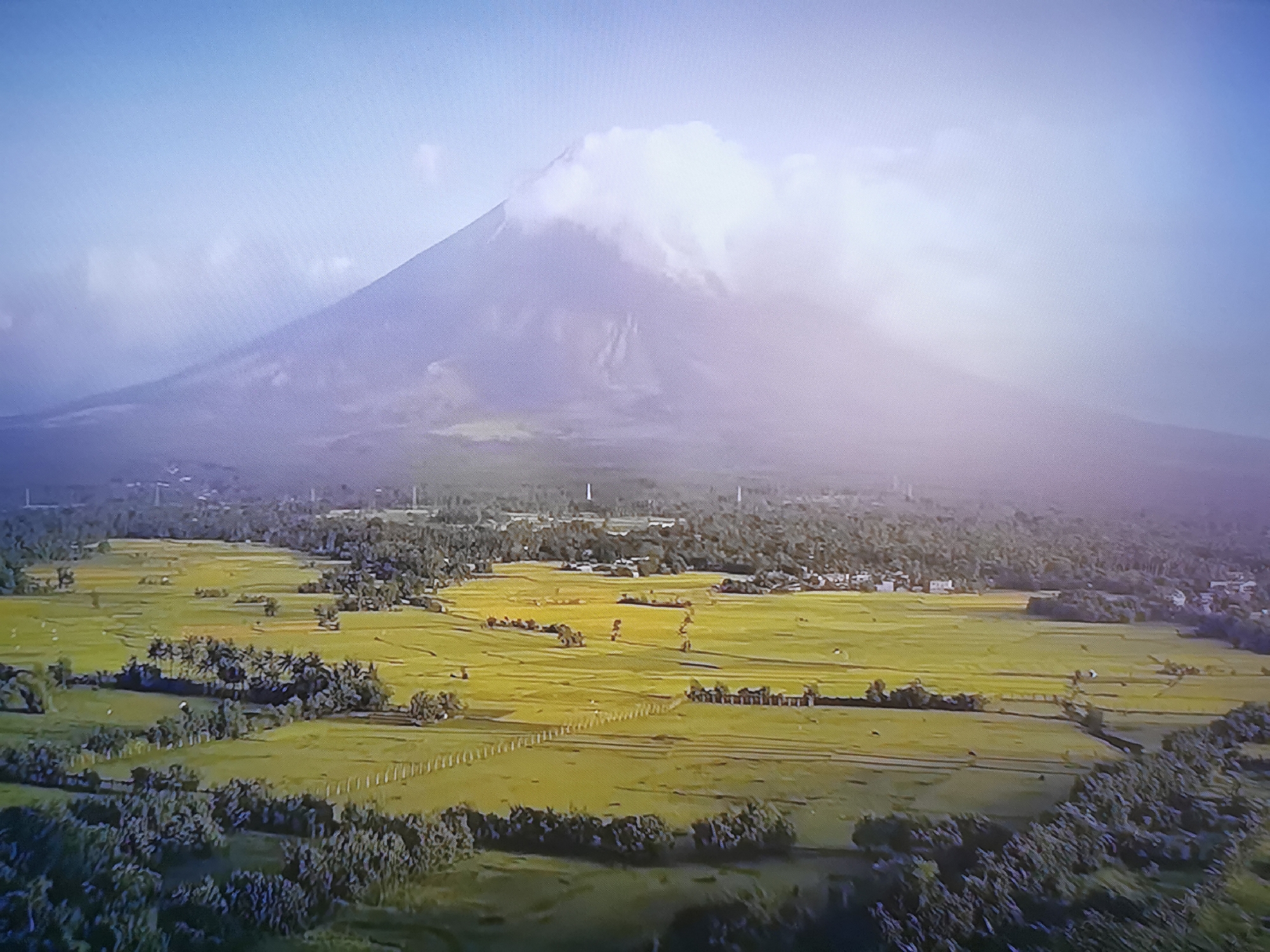 The image is an aerial view of a volcano with a cloud cap, set against a bright blue sky. In the foreground, there are vast, verdant fields, likely agricultural land, with scattered trees and some evidence of human settlements or structures. The fields appear to be irrigated or are a result of recent rainfall, giving them a lush green appearance. The overall scene depicts a serene natural landscape with a prominent volcano in the background. The location is not in the city of Dnipro, Ukraine, but rather a natural volcanic landscape.