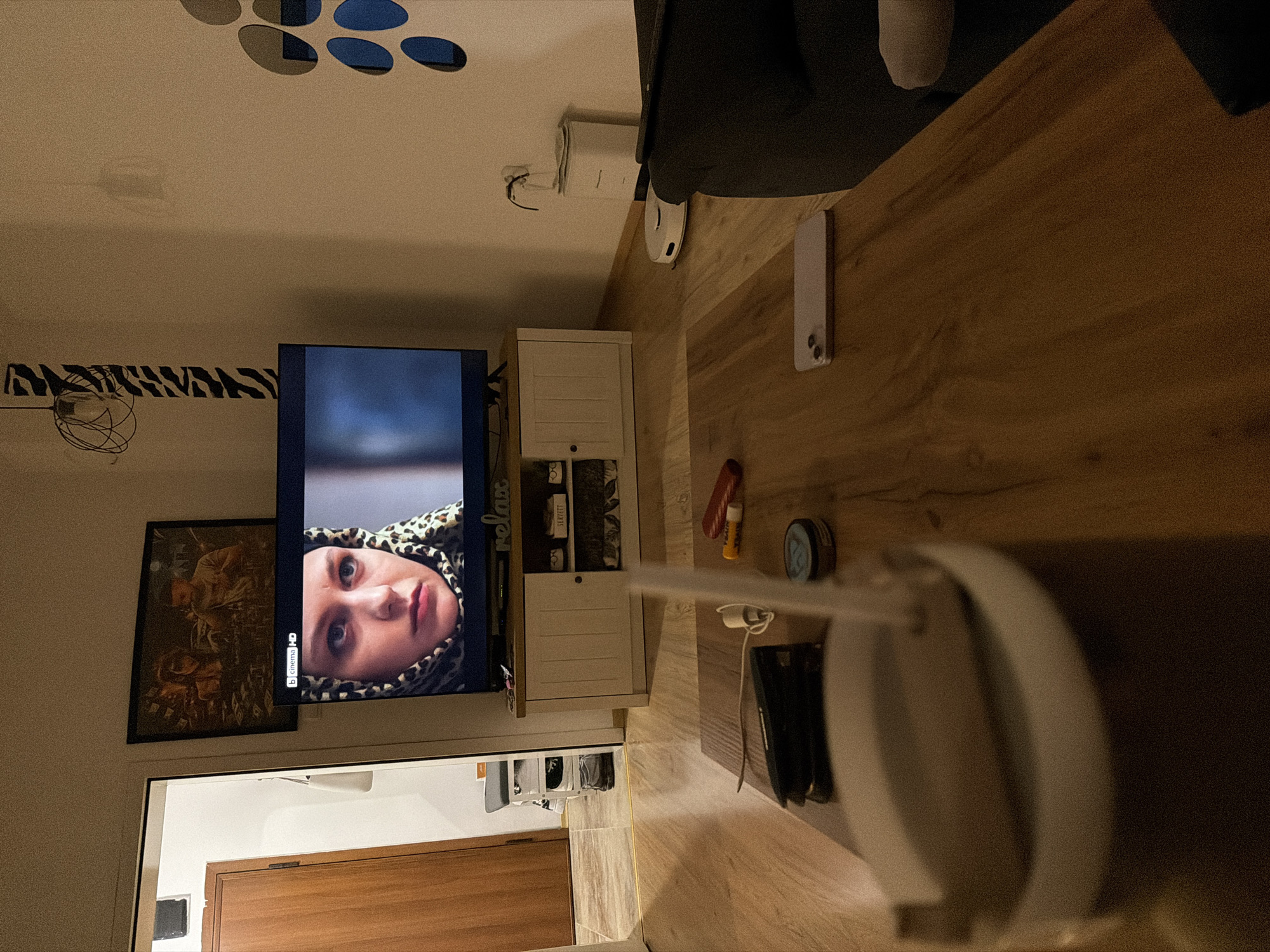 The image depicts an indoor scene, likely a living room, with the focus on a television screen displaying a close-up of a woman's face. To the left of the television, a wooden door is visible, suggesting an entryway. The television is placed on a white cabinet, which has several compartments. On the cabinet, a framed picture hangs on the wall above, and a decorative word "relax" is also visible. To the right of the television, a white robot vacuum cleaner is present on the floor, near a white appliance. The foreground is dominated by a wooden coffee table, upon which several objects are scattered: a stapler, a lip balm, a small blue tin, a white cup with a straw, and a smartphone. The overall lighting suggests it is evening or a dimly lit interior. There is no information to determine the location within the city of Plovdiv, Bulgaria.