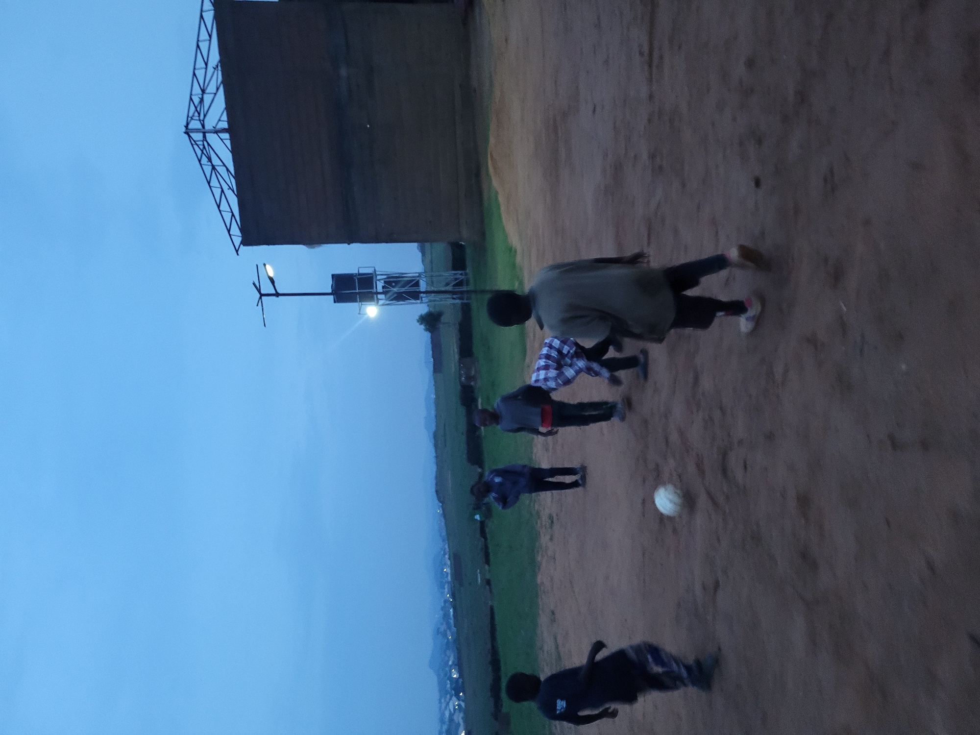 In the city of Barakin Akawo, Nigeria, at dusk, five young boys are playing soccer on a dirt field. A large, dark brown building with a metal frame roof structure is visible to the right of the frame. To the left of the building, a tall water tower with lights is illuminated. The sky is a deep blue, indicating the transition from day to night. The ground is a mix of sand and dirt, with some scattered debris. One boy on the far left is wearing a dark t-shirt and patterned pants, actively kicking a soccer ball. Another boy in the center is wearing a grey shirt and dark pants, leaning forward as if reaching for the ball. To the right of him, a boy in a blue and grey checkered shirt and dark pants stands looking at the ball. Further back, two other boys are also present, their actions and positions suggesting their involvement in the game. The environment appears to be on the outskirts of a town or city, with rolling hills and scattered lights visible in the distance.