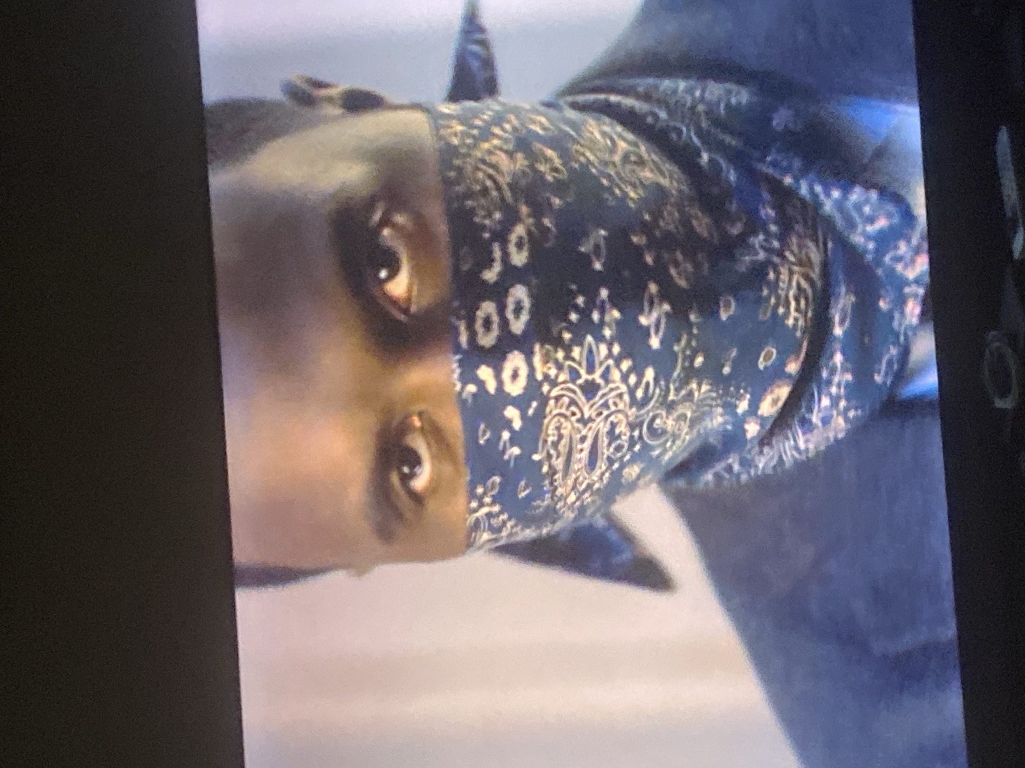 The rotated image captures a close-up of a person, focusing on their face from the nose upwards. The individual has dark skin and is looking directly at the viewer. They are wearing a dark blue bandana with a light-colored paisley pattern, which covers the lower part of their face and forehead. Part of a dark blue garment is visible around their shoulders. The background is blurred, showing a light-colored surface below and a darker, bluish-grey area above, suggesting an indoor setting. The image quality is grainy, and a dark border is present on the left side, indicating it might be a photograph of a screen or a screenshot. No specific activity or event is discernible beyond the person's attentive gaze. There are no visible texts, and no specific time of day or weather conditions can be determined from the visual cues. The image itself does not provide any specific location cues related to Abuja, Nigeria.
