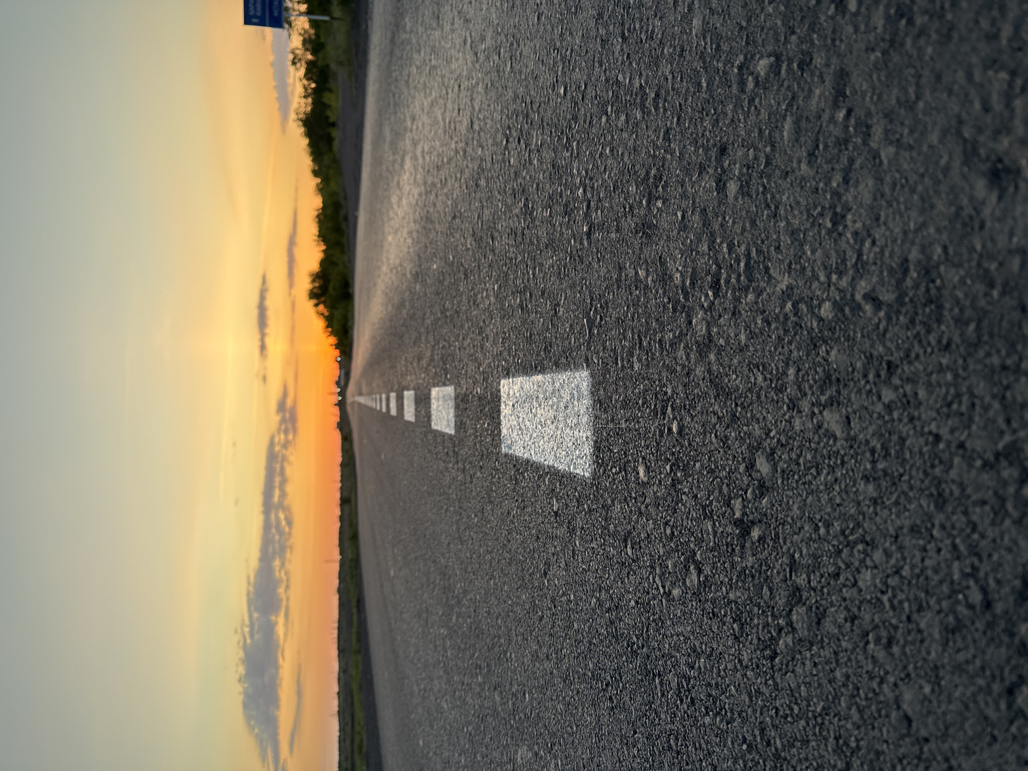 The image shows an asphalt road stretching into the distance under a sky with a vivid sunset. The road is marked with a series of rectangular white lane dividers down its center. The texture of the asphalt is coarse and detailed. On the left side of the road, a colorful sunset with hues of yellow, orange, and hints of blue can be seen. The location is Unknown, Unknown.