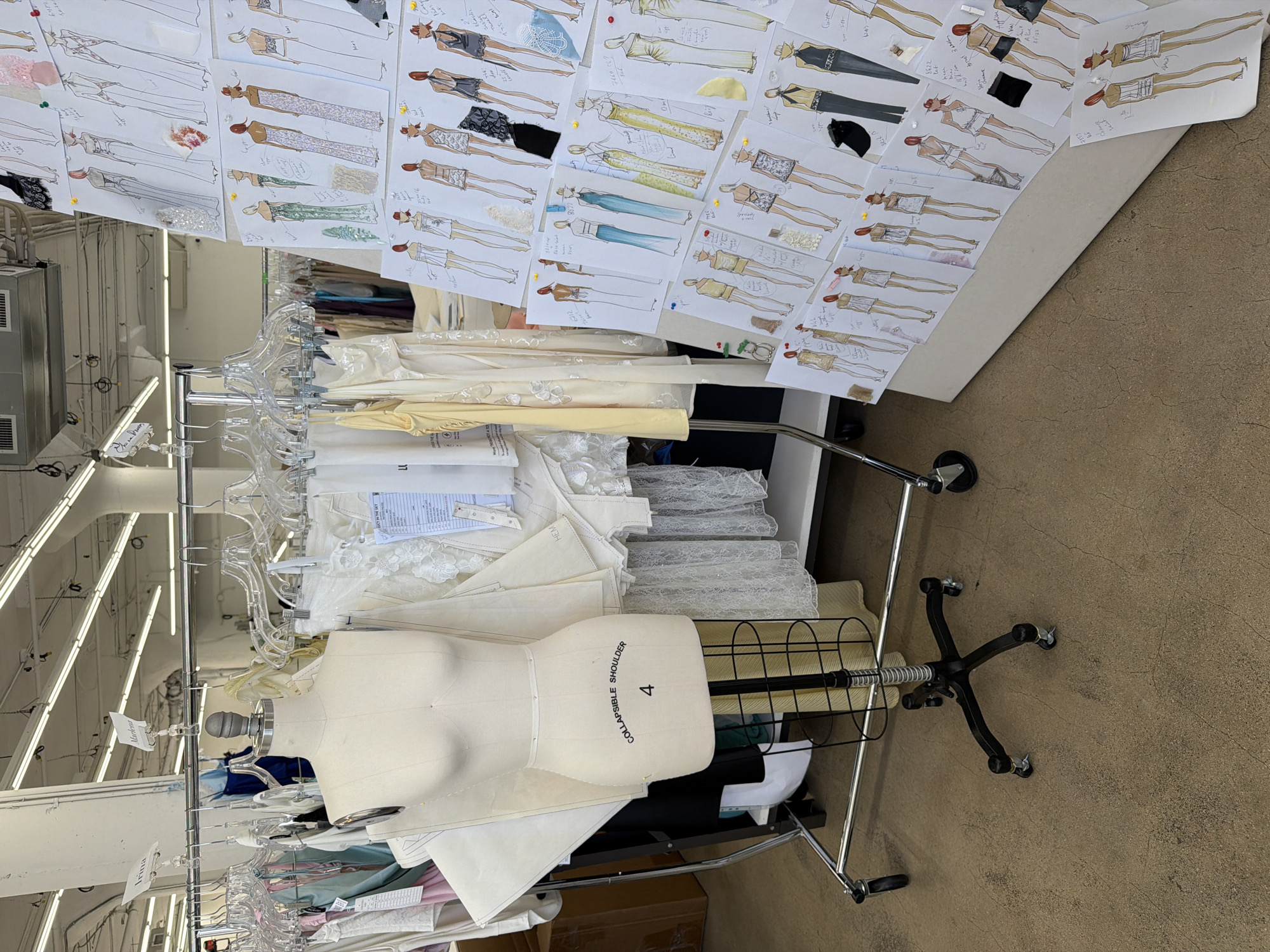 The image shows a sewing studio in Los Angeles, California. The studio is filled with garment racks, a dress form, and sketches of dresses. The garment racks are filled with various fabrics and unfinished garments. The dress form is a female mannequin that is a size 4. The sketches are of dresses in various styles and colors. There are also some fabric swatches attached to the sketches. The room is lit by fluorescent lights and there is a concrete floor. The overall impression is one of a busy and creative workspace.