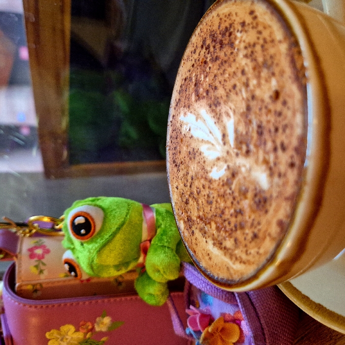 An indoor scene, likely a cafe in Strasbourg, France, is captured in a close-up perspective. In the foreground, a light brown ceramic cup holds a frothy beverage, presumed to be coffee, topped with cocoa powder and a white, leaf-shaped latte art design. The cup rests on a partially visible, matching saucer. To the left, a bright green plush chameleon toy, featuring large black-and-white eyes and a pink ribbon around its neck, is suspended by a gold-colored clip. Behind the toy, a magenta bag with yellow, orange, and pink floral patterns is partially visible. Adjacent to the bag, a small purple basket-like object containing miniature colorful flowers is also present. The background on the right displays a dark, reflective surface, consistent with a window or glass pane, framed by dark wood. Blurred green and blue reflections are discernible within the glass. No specific identifying landmarks are present within the image to pinpoint the exact location within Strasbourg.