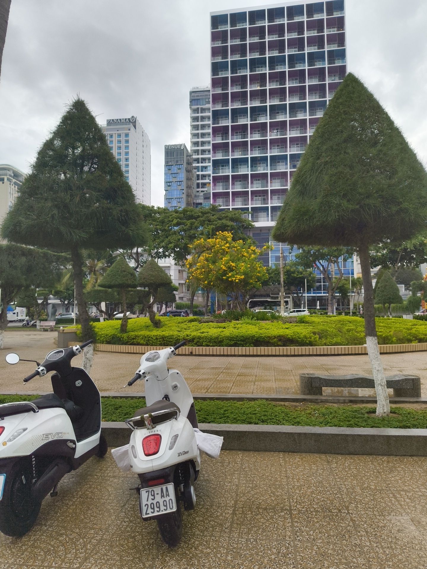 Two white electric scooters are parked on a patterned paved area in Nha Trang, Vietnam. The scooter on the left is black and white with "EVO" visible on its side. The scooter on the right is white with a brown seat and a visible license plate reading "79-AA 299.90". Behind the scooters, a neatly manicured green circular garden bed is surrounded by a low stone wall. Tall, conical trees flank the garden. In the background, modern high-rise buildings with balconies are visible, including a building with a grid pattern of blue and purple square openings. The sky is overcast, suggesting a cloudy day. The pavement appears wet, indicating recent rain.