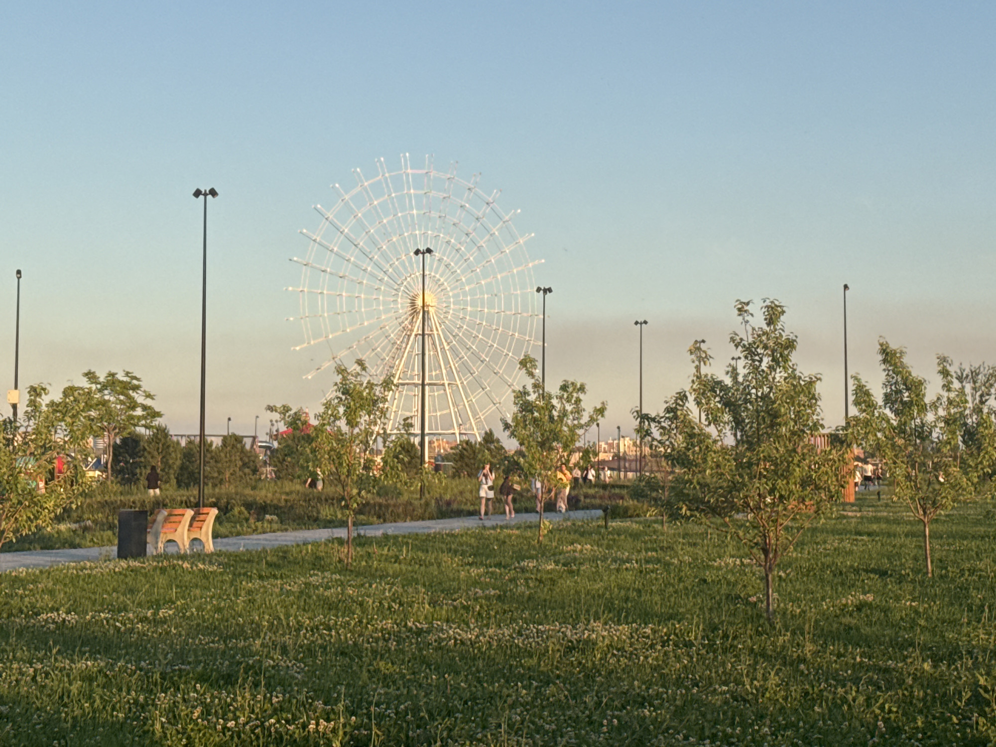 The image depicts a Ferris wheel in the background, located in the city of Magnitogorsk, Russia. In the foreground, a park area is visible with a path and several trees. The grass is green and sparsely dotted with small white flowers. A park bench with a trash can next to it is situated on the left side of the path. Several people are walking along the path and in the background. The sky is clear and blue, with the sun setting, casting a warm glow on the scene.