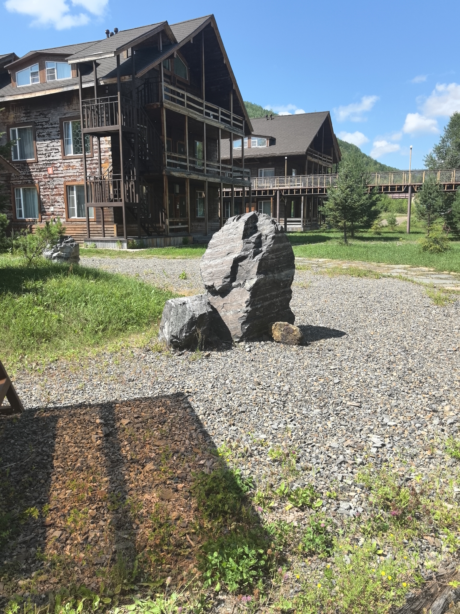 The image depicts the exterior of a multi-story wooden lodge-style building, possibly a hotel or resort, with a weathered appearance. The building has multiple balconies and a visible exterior staircase. In the foreground, a large, dark, layered rock is prominently featured. The ground is covered with gravel, and there is a patch of green grass to the left of the rock. A wooden structure, possibly a bench or part of a shelter, casts a shadow on the gravel in the lower left of the frame. In the background, another similar building is visible, connected by an elevated walkway or bridge. The scene is set under a clear blue sky with scattered white clouds. The environment appears to be a natural setting, possibly mountainous or wooded, given the presence of trees and a distant hill. This location is identified as being in the city of Solnechnyi, Russia. There are no people or animals visible in the image.